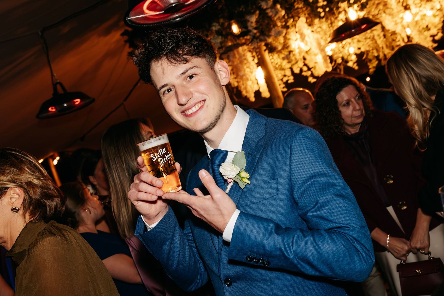 Young man in blue suit smiling and holding a beer at a wedding reception.