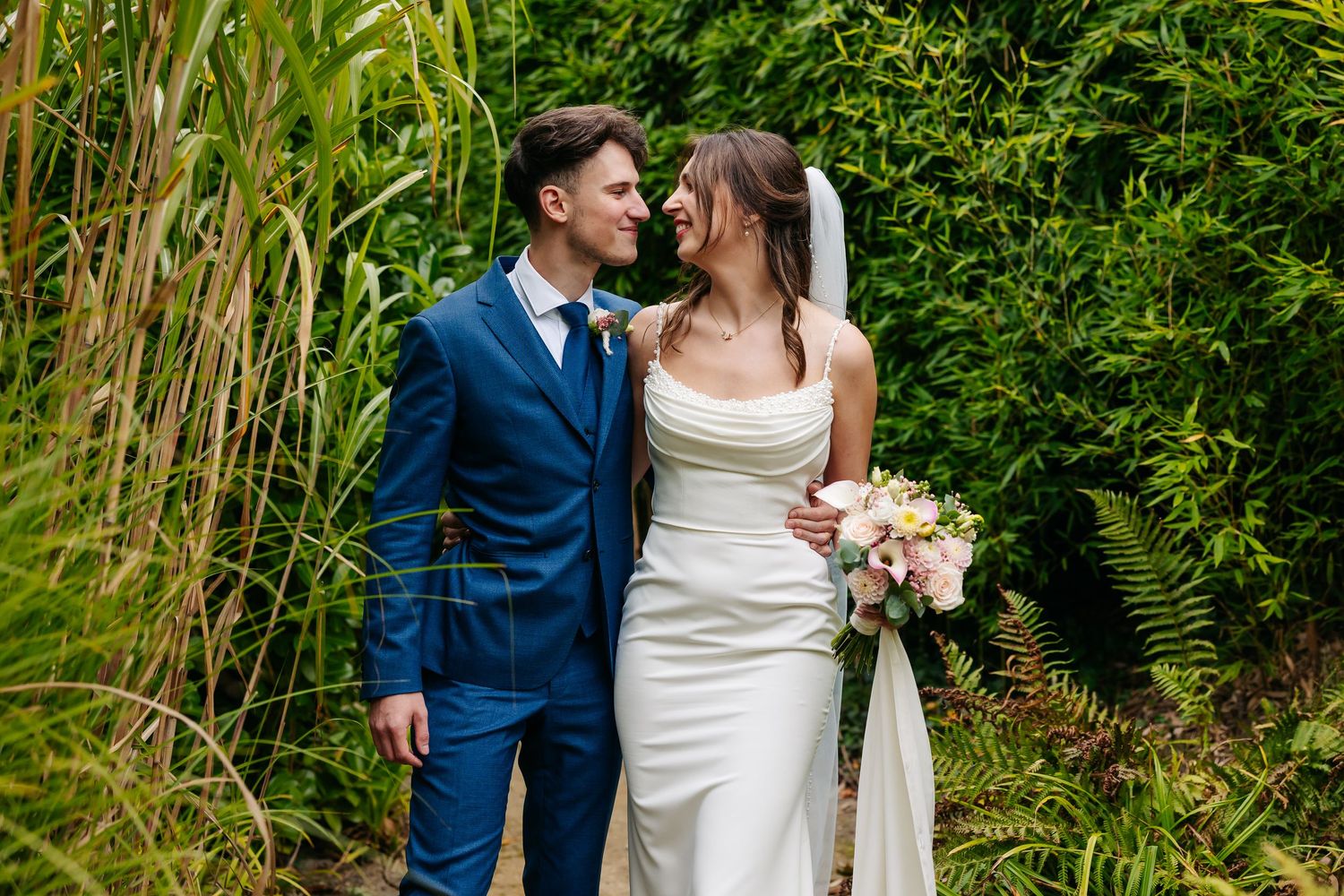 Bride and groom gazing at each other romantically surrounded by tall green plants outdoors.