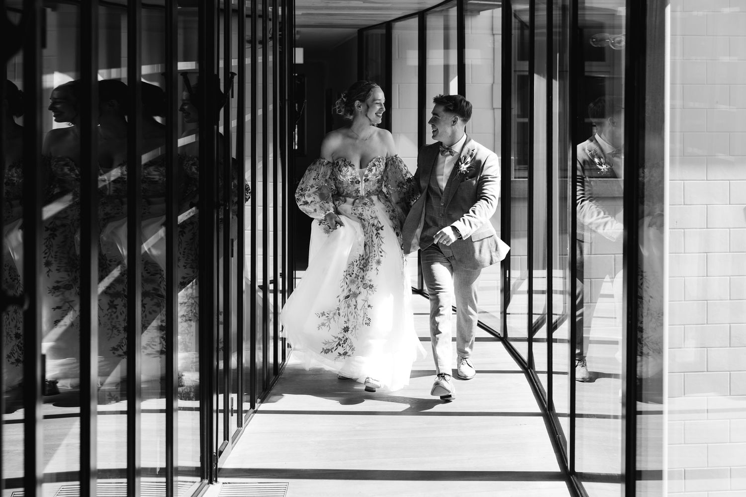 Black and white photo of bride and groom walking through modern architectural corridor on wedding day.