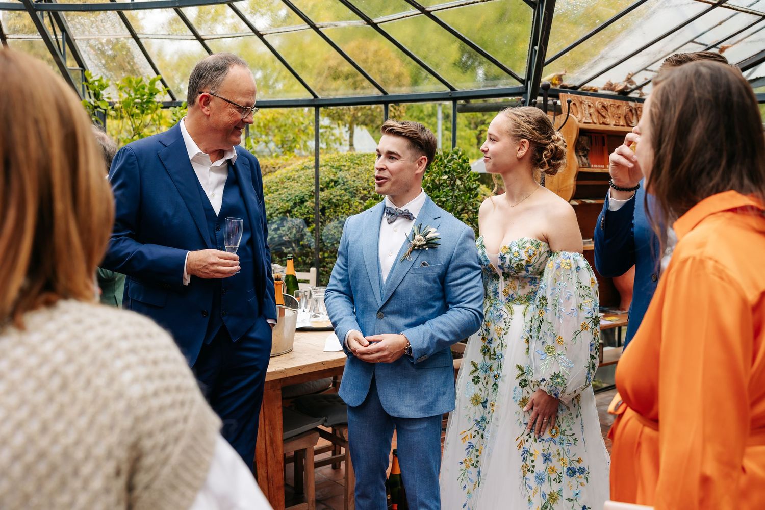 Bride and groom in floral dress and blue suit socializing with wedding guests inside a greenhouse venue.