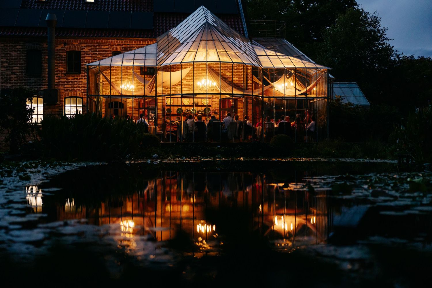 Glowing glass pavilion wedding venue reflected in a lily pond at dusk.