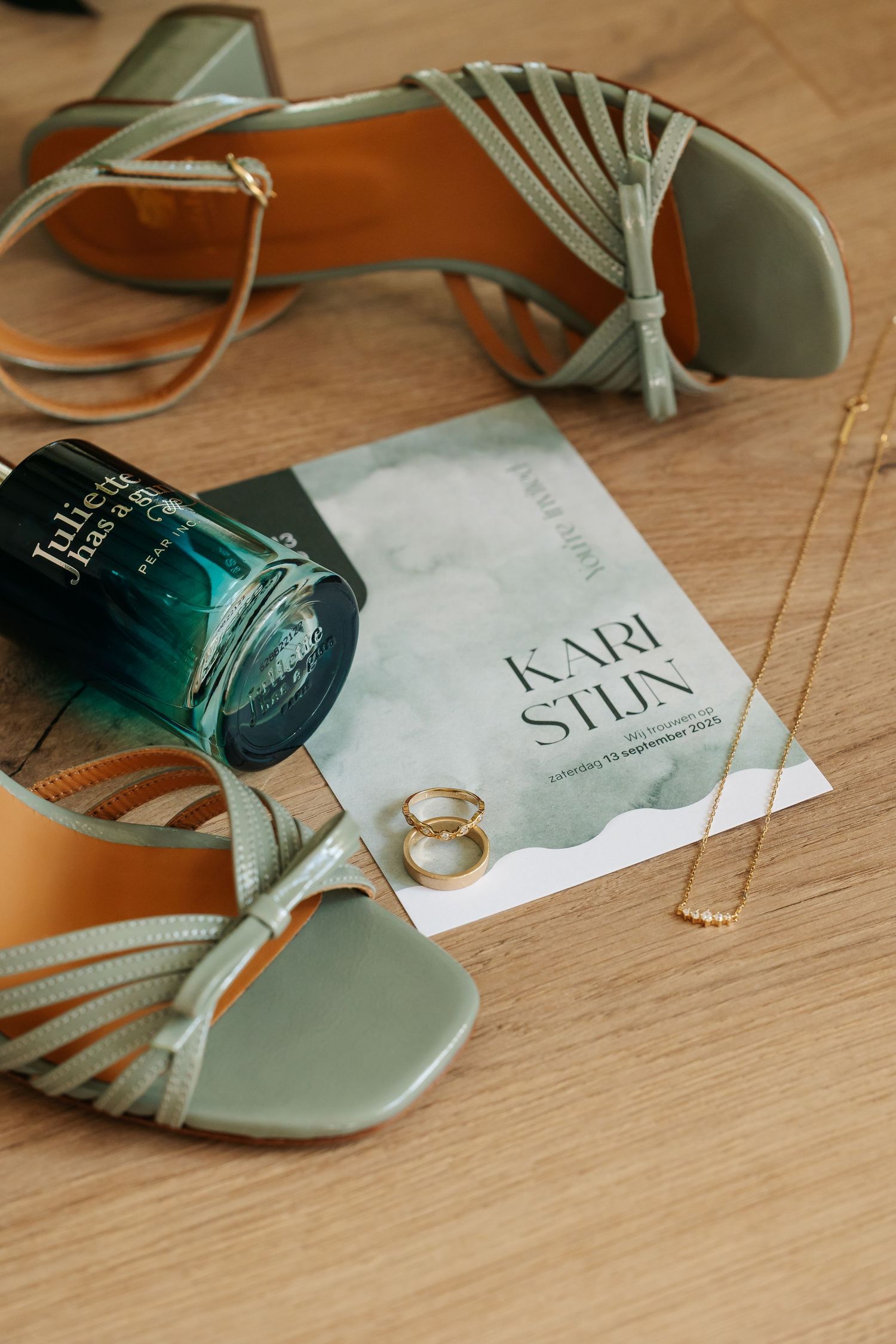 Sage green wedding heels, perfume, gold necklace, rings, and invitation styled on wood floor.