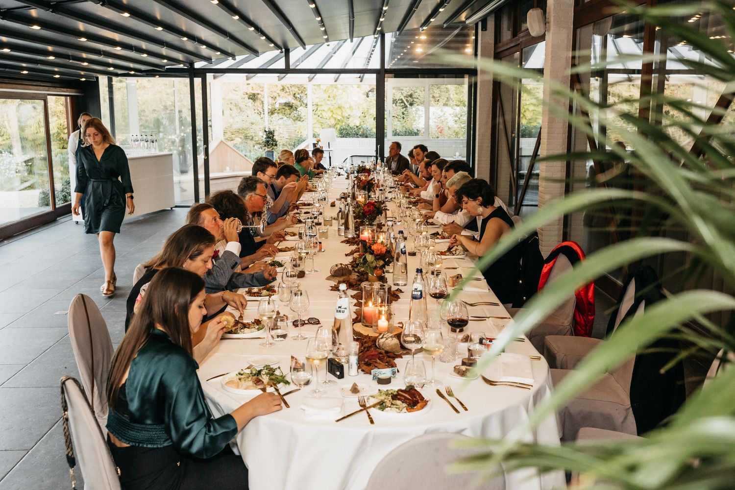 Guests seated at a long elegantly set dining table inside a modern glass-walled event venue.