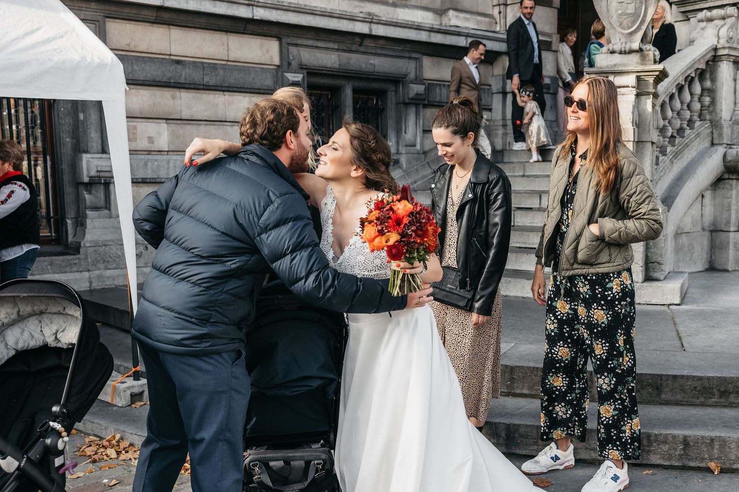 Bride in white gown with autumn bouquet receiving congratulations from guests on city street.