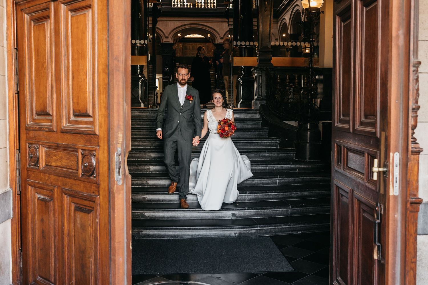 Bride and groom descend grand marble staircase through ornate wooden doors at elegant wedding venue.