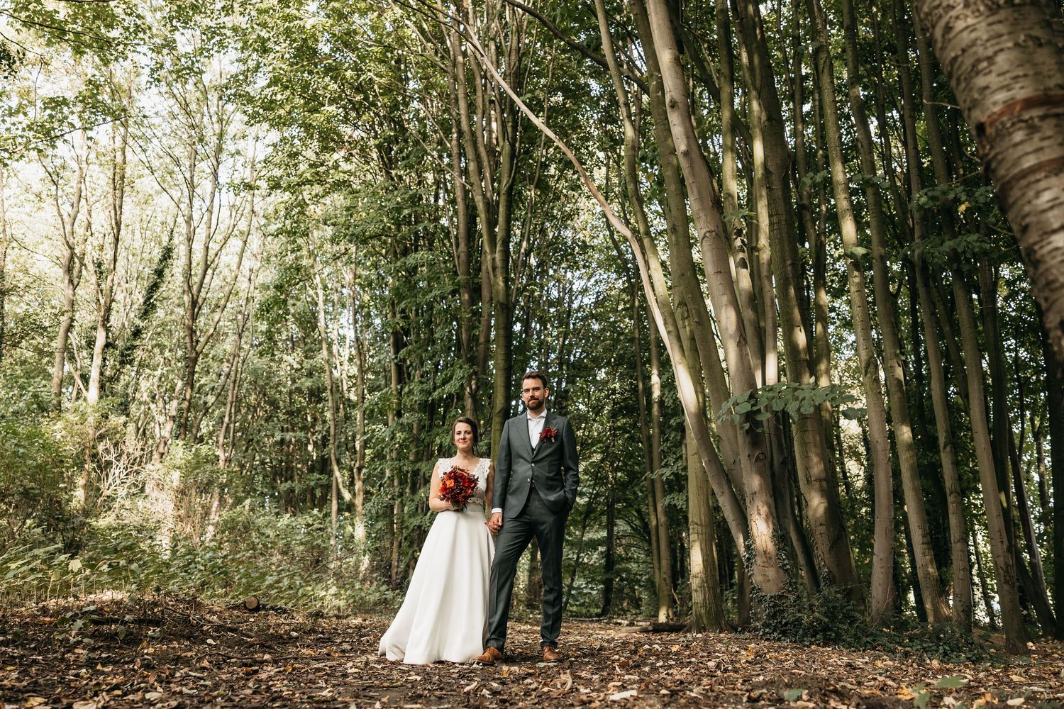 Bride and groom standing apart in tall forest trees on their autumn wedding day.