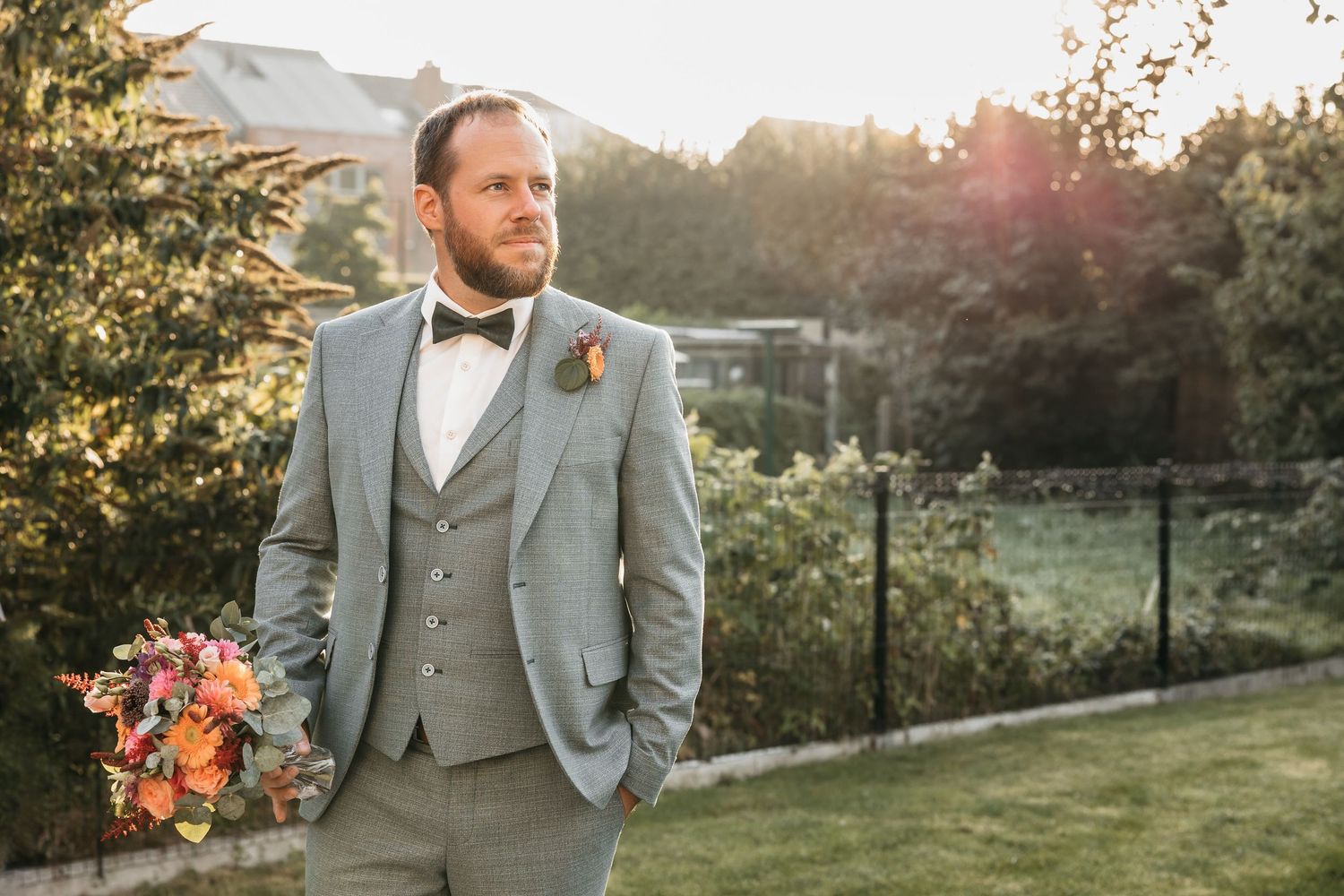Groom in grey three-piece suit holding a vibrant bouquet in a sunlit garden setting.