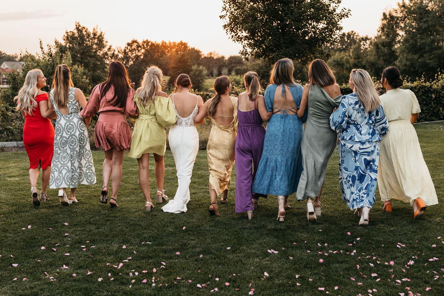 Bride and female guests in colorful dresses walking away together at outdoor sunset wedding.