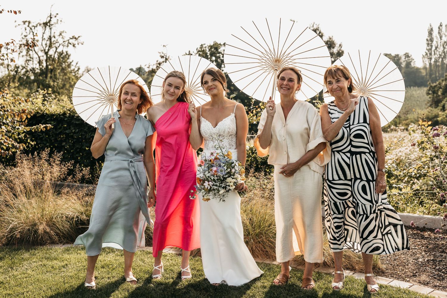 Laughing bride with bridesmaids in colorful dresses holding white parasols on a sunny wedding day.