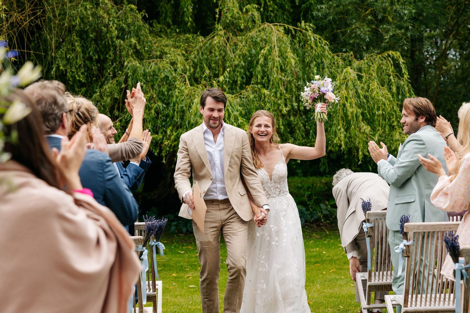 Newlyweds walk joyfully down outdoor aisle as guests cheer, bride in lace gown holding bouquet, groom in tan suit.