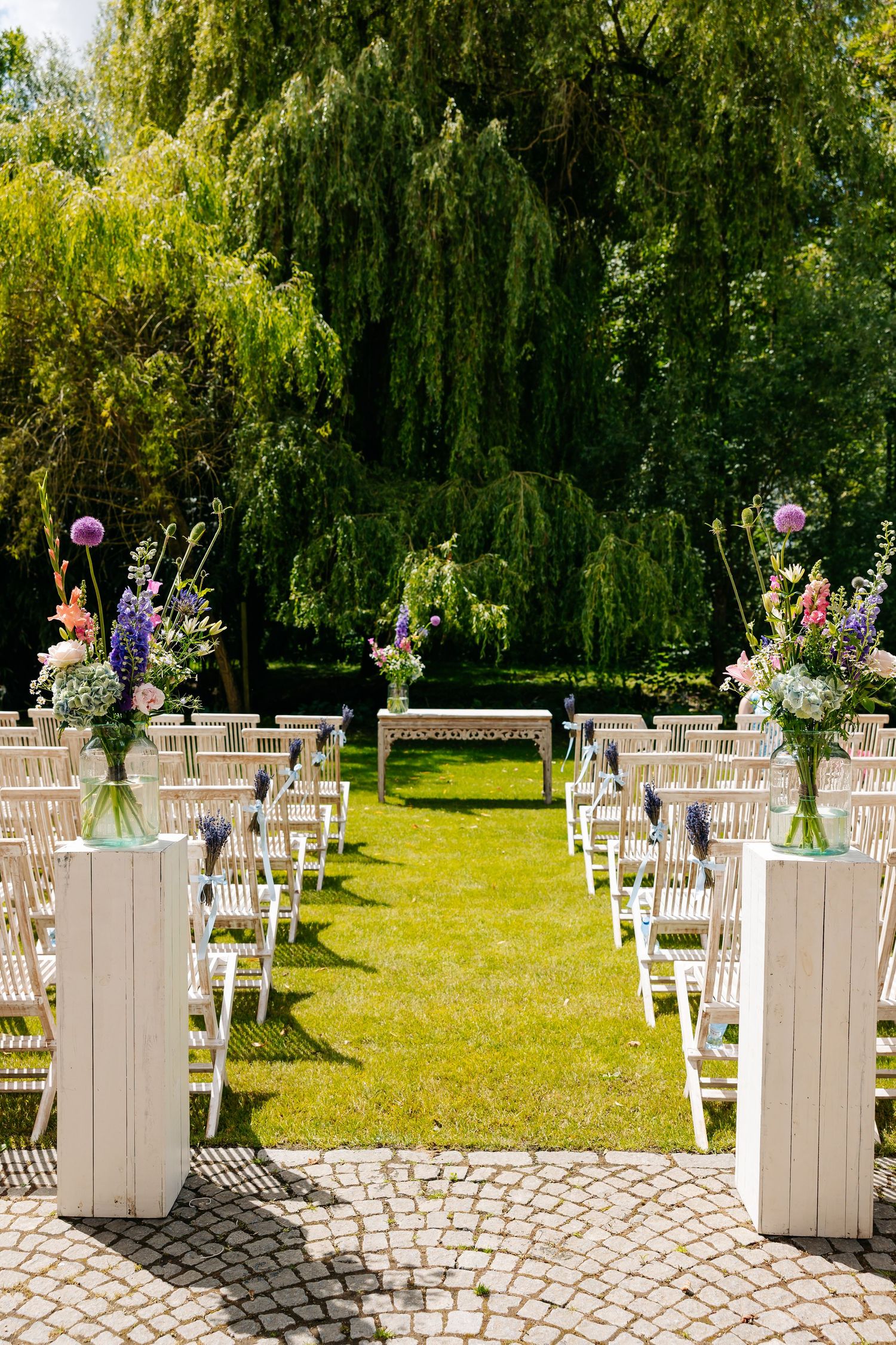 Elegant outdoor wedding ceremony setup with white chairs, floral arrangements, and a lush green garden backdrop.