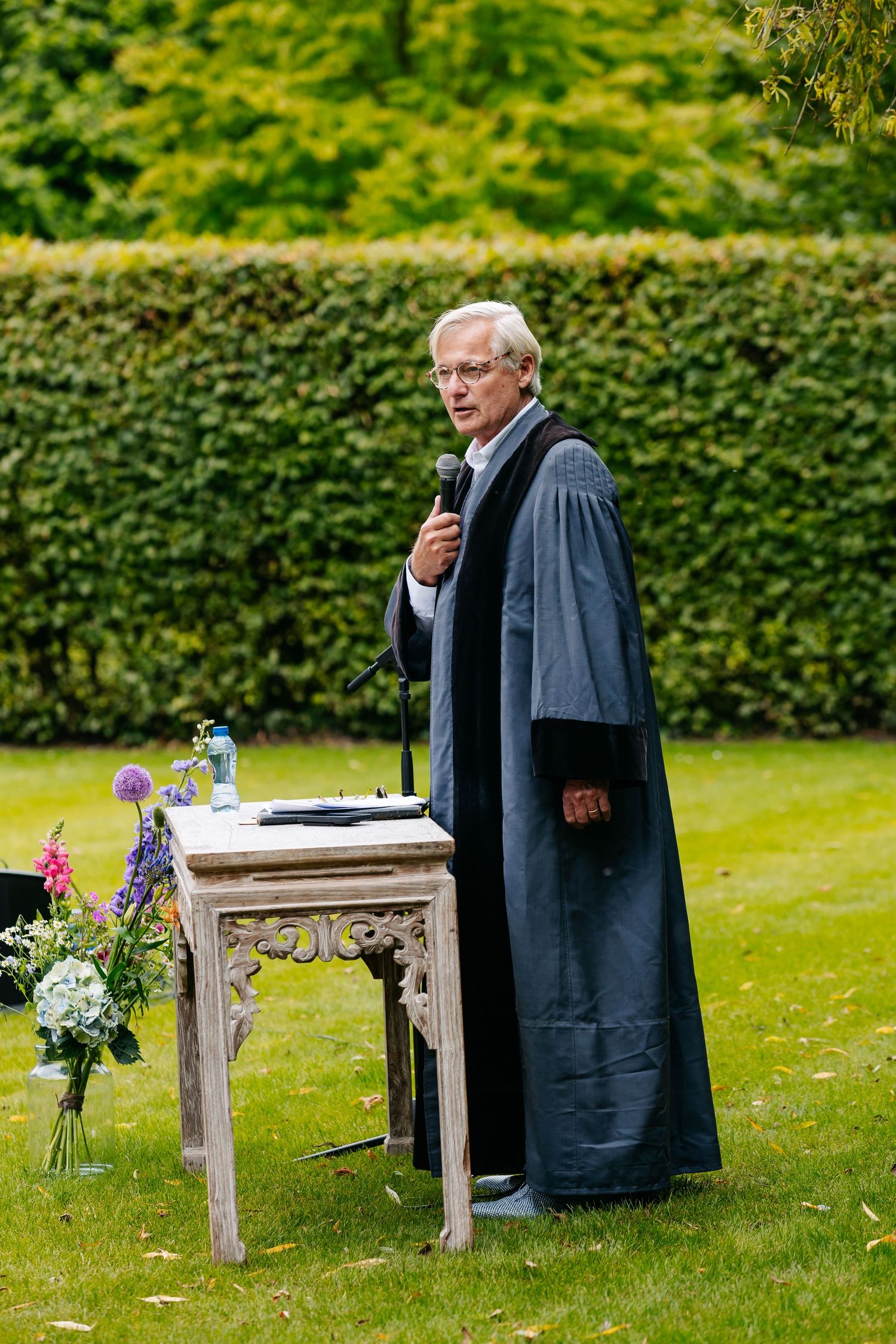 Officiant in gray robes speaks at an ornate outdoor wedding ceremony altar surrounded by greenery.