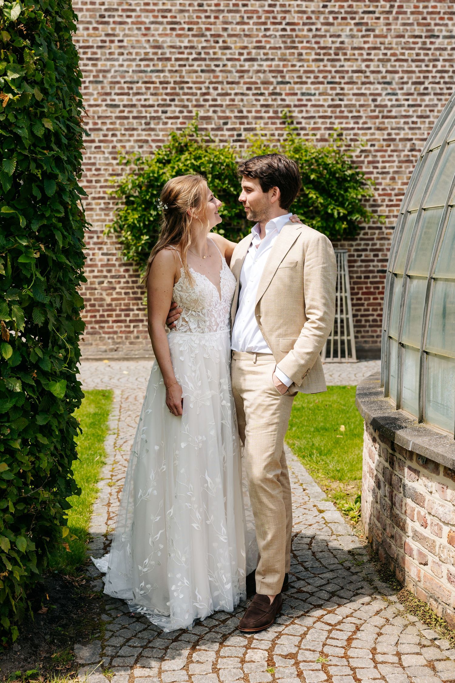 Bride in lace gown and groom in beige suit pose together in a garden courtyard with brick wall backdrop.