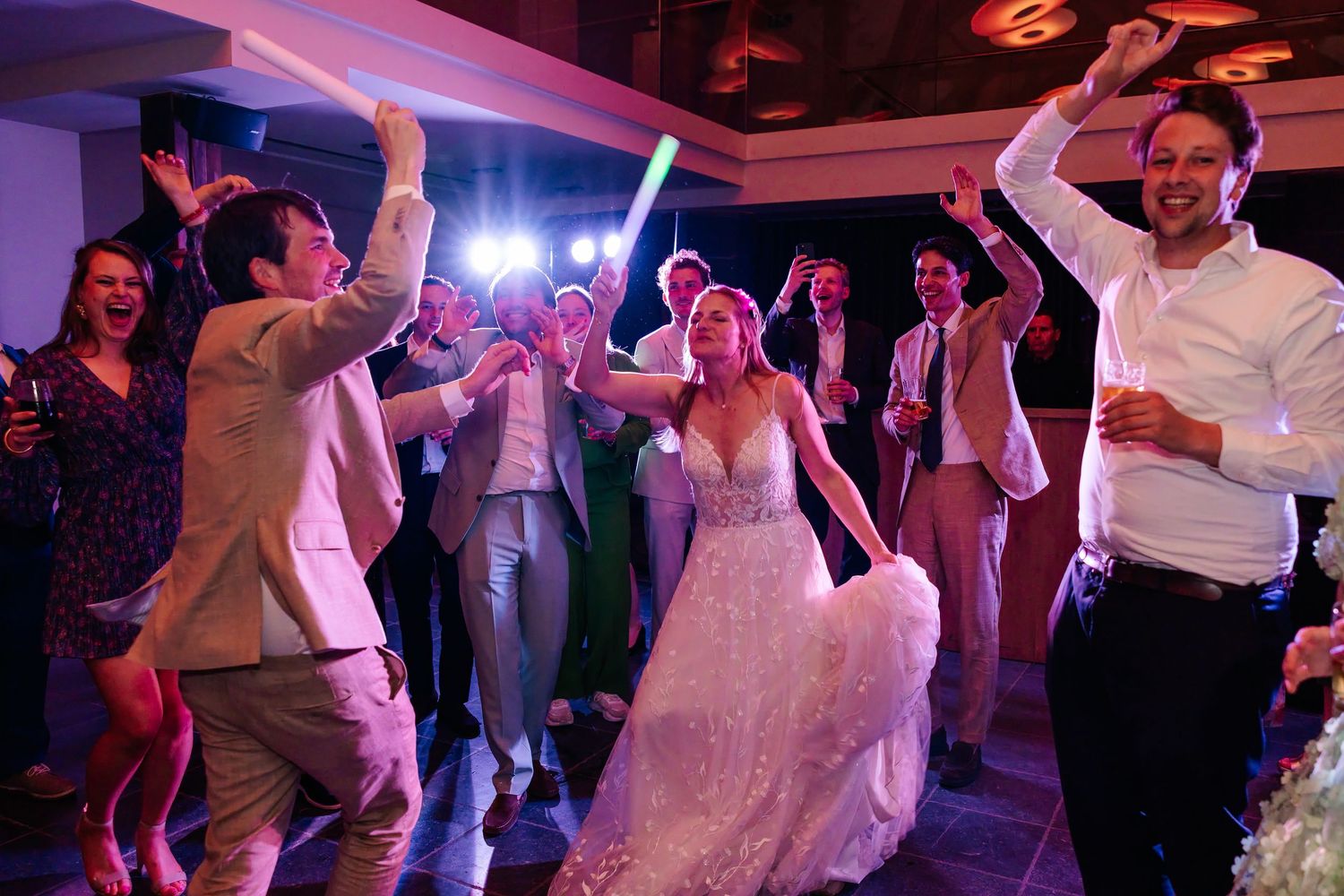 Bride in lace gown dancing joyfully with groom and guests under colorful lights at wedding reception.