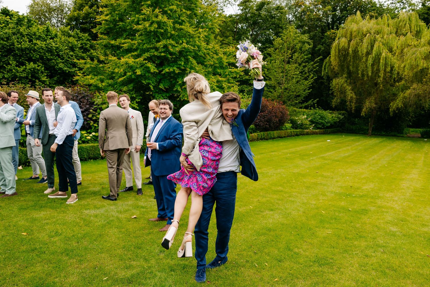 Excited wedding guest lifts woman in pink skirt while holding bouquet on green lawn at reception.