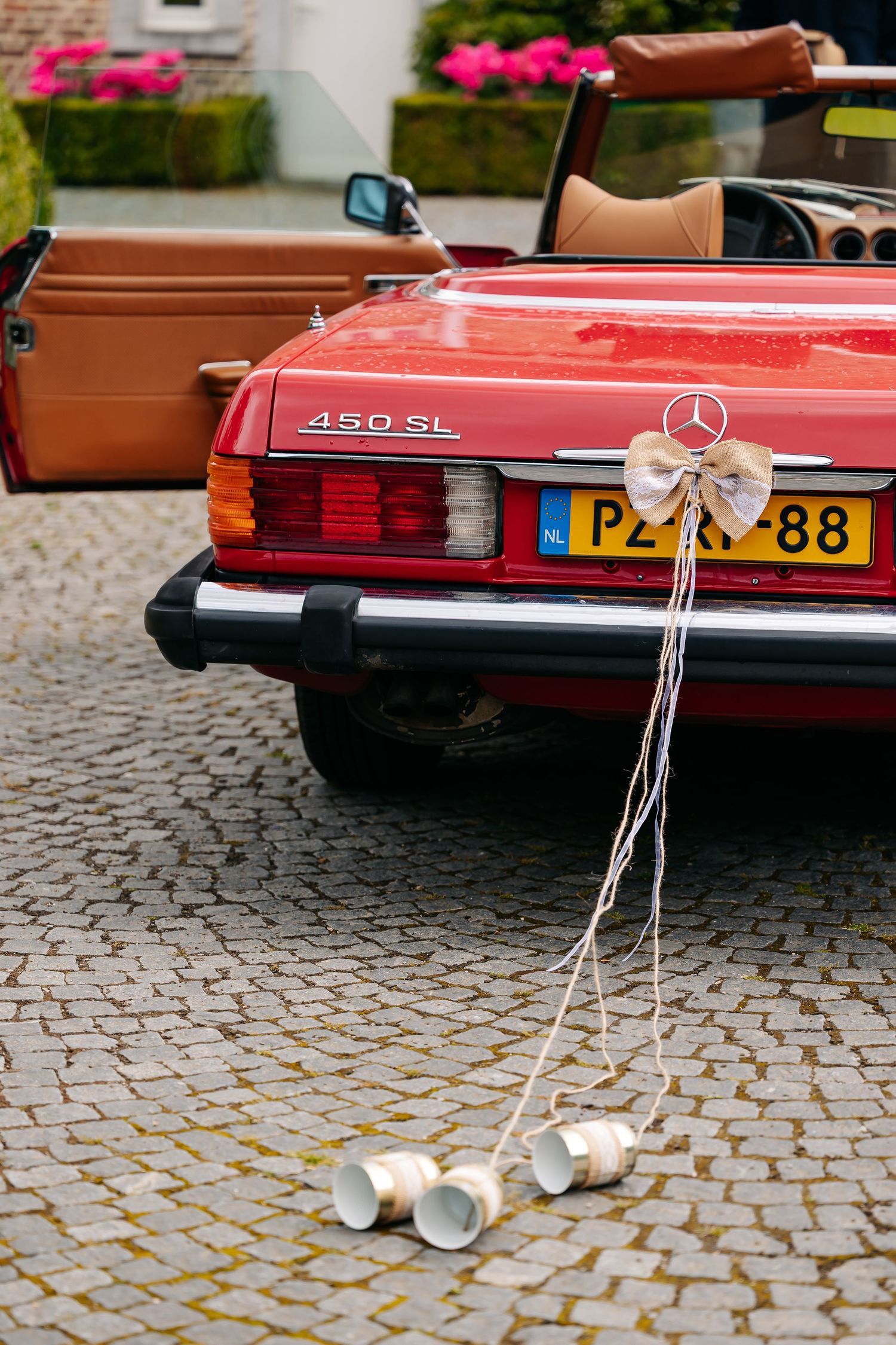 Red Mercedes-Benz 450SL convertible wedding car with tin cans tied to bumper and bow decoration on cobblestone driveway.