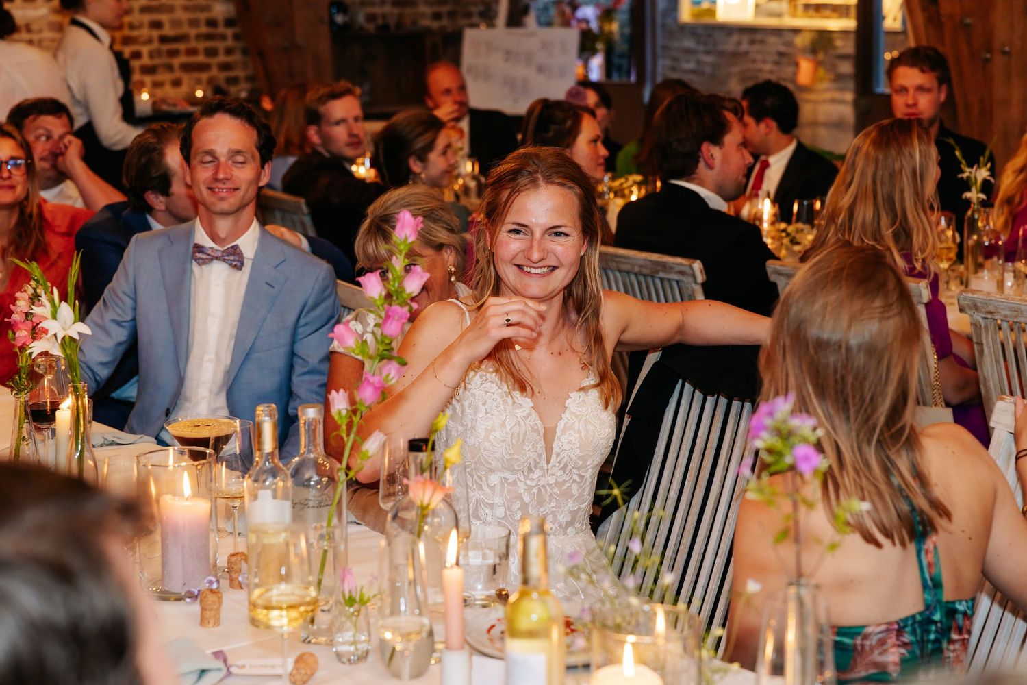 Smiling bride in a lace gown seated at a candlelit wedding reception dinner table with guests.