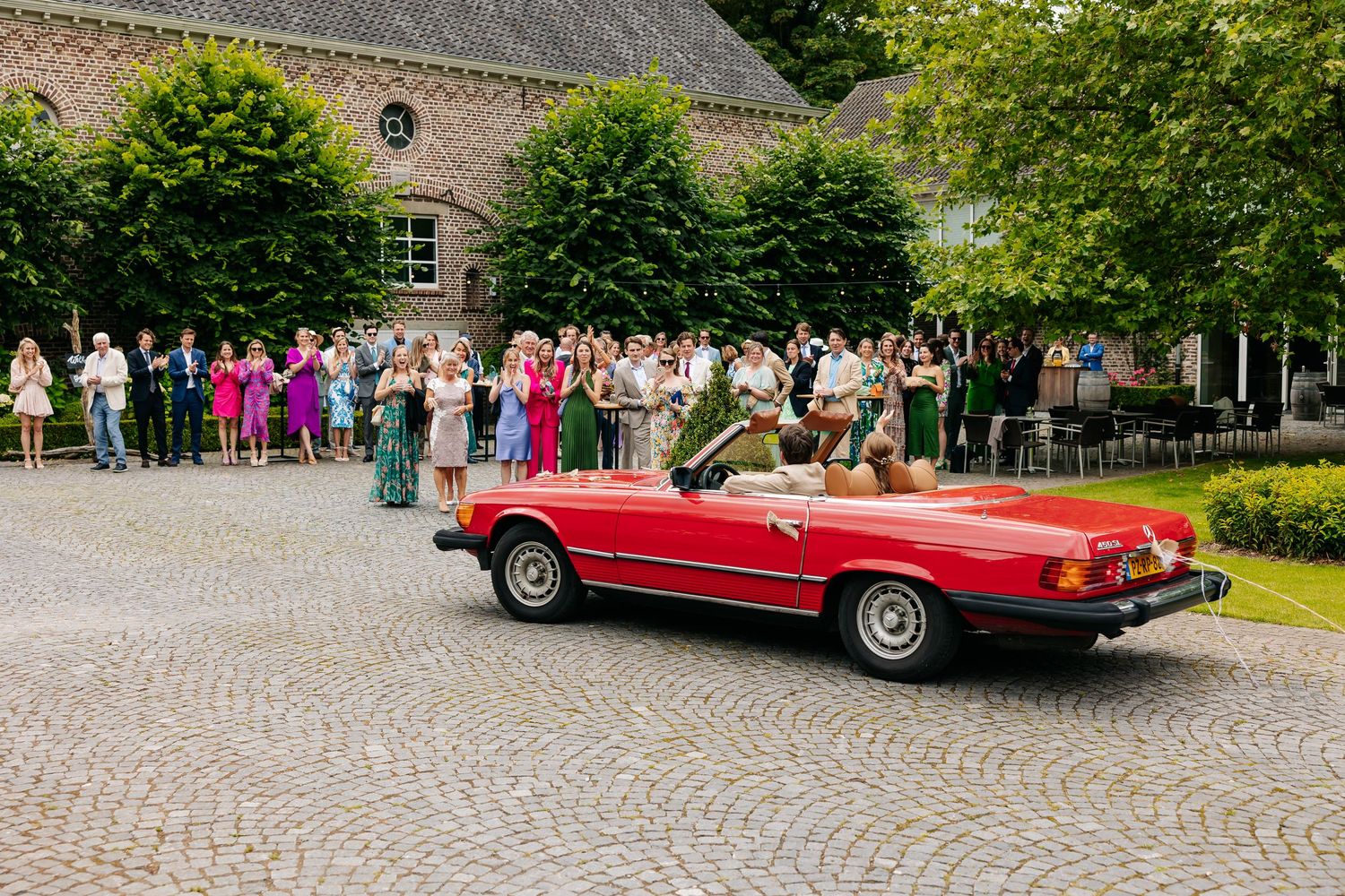 Red vintage convertible drives past cheering wedding guests on a cobblestone venue.