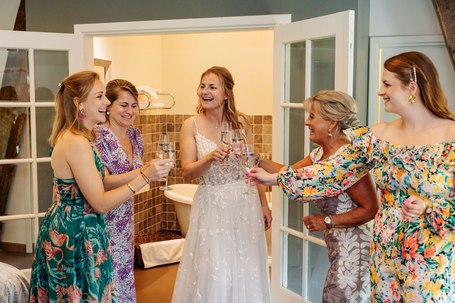 Bride in wedding dress laughing and toasting champagne with four bridesmaids in floral dresses.