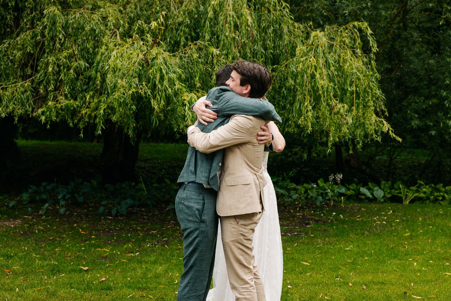 Two men share a heartfelt hug in a lush green garden during a wedding celebration.