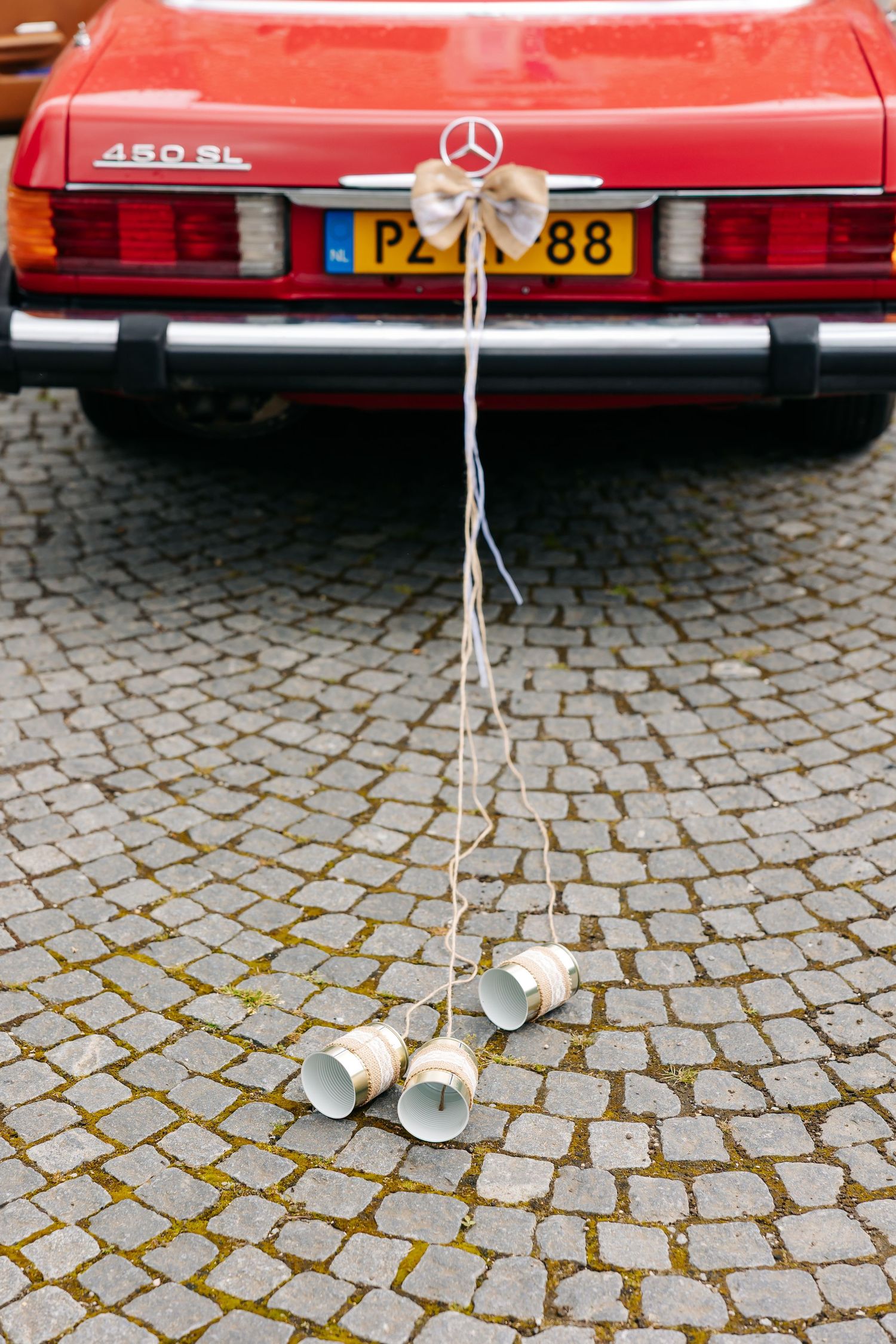 Red Mercedes-Benz 450SL wedding car with tin cans tied to bumper on cobblestone street.