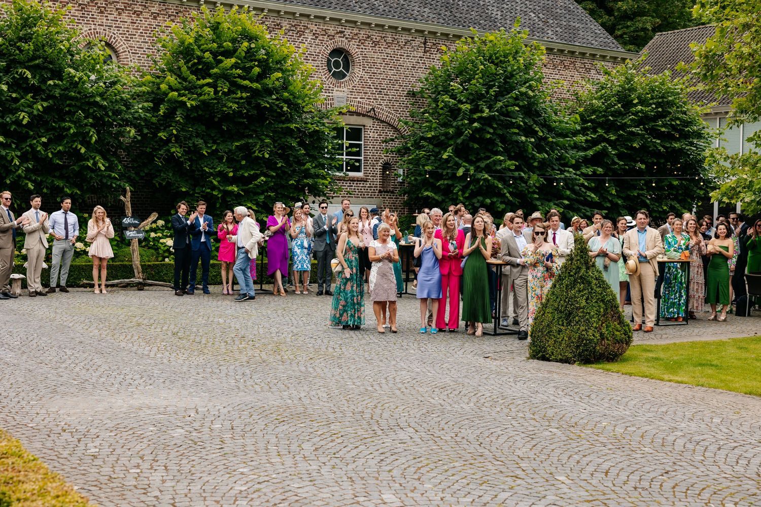 Large wedding guest crowd gathered on cobblestone courtyard outside rustic brick estate venue.