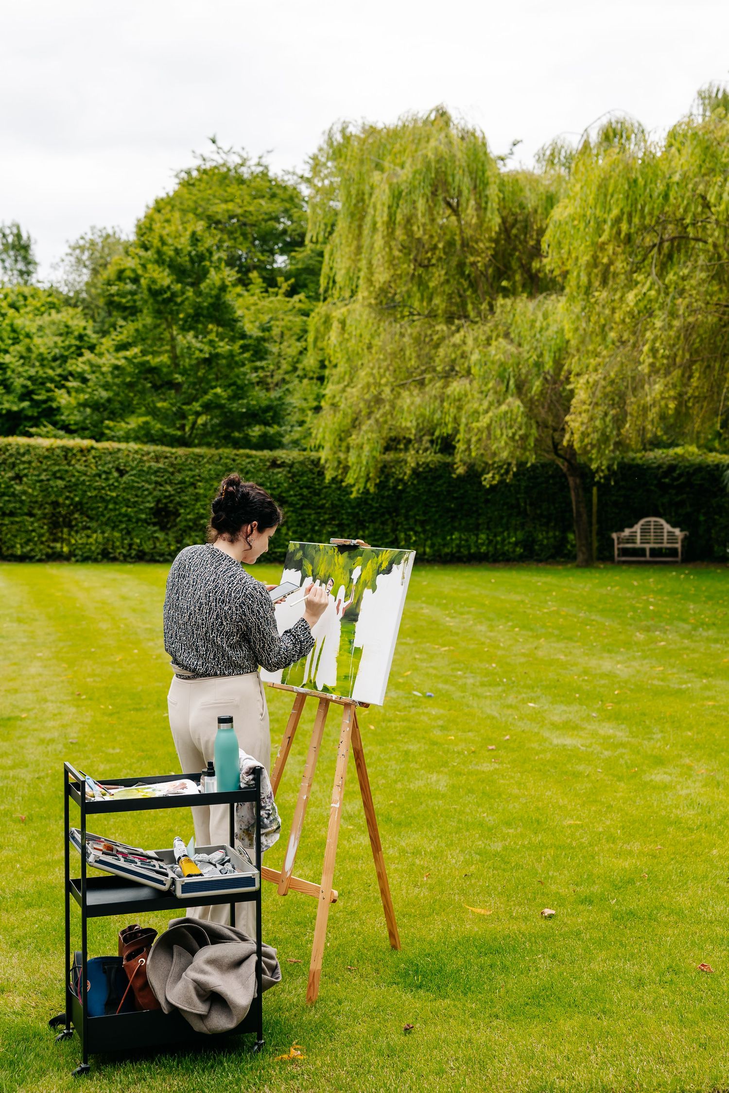 A woman paints on an easel outdoors in a lush green garden at a wedding event.