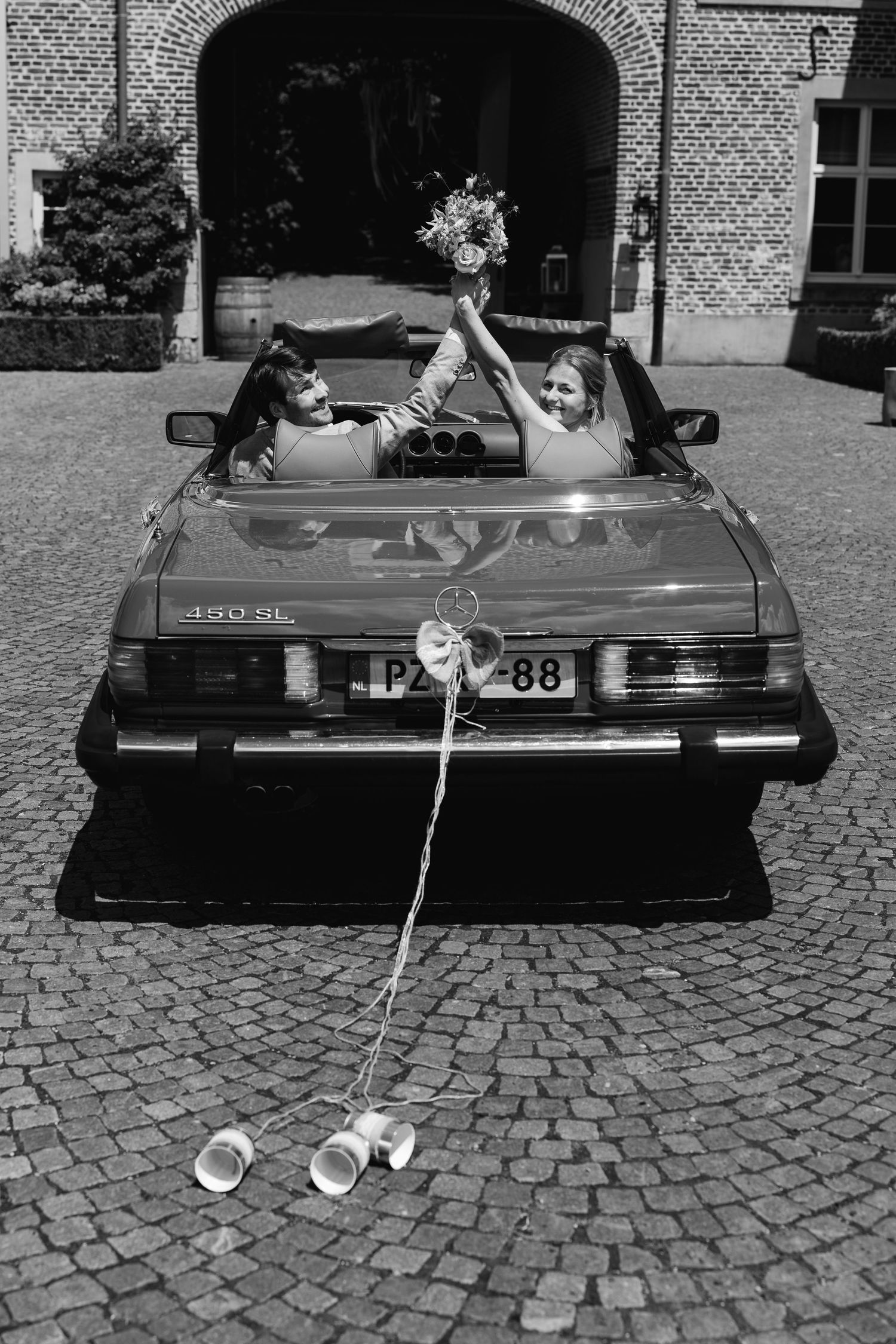Black and white photo of newlyweds celebrating in vintage convertible car decorated with tin cans.