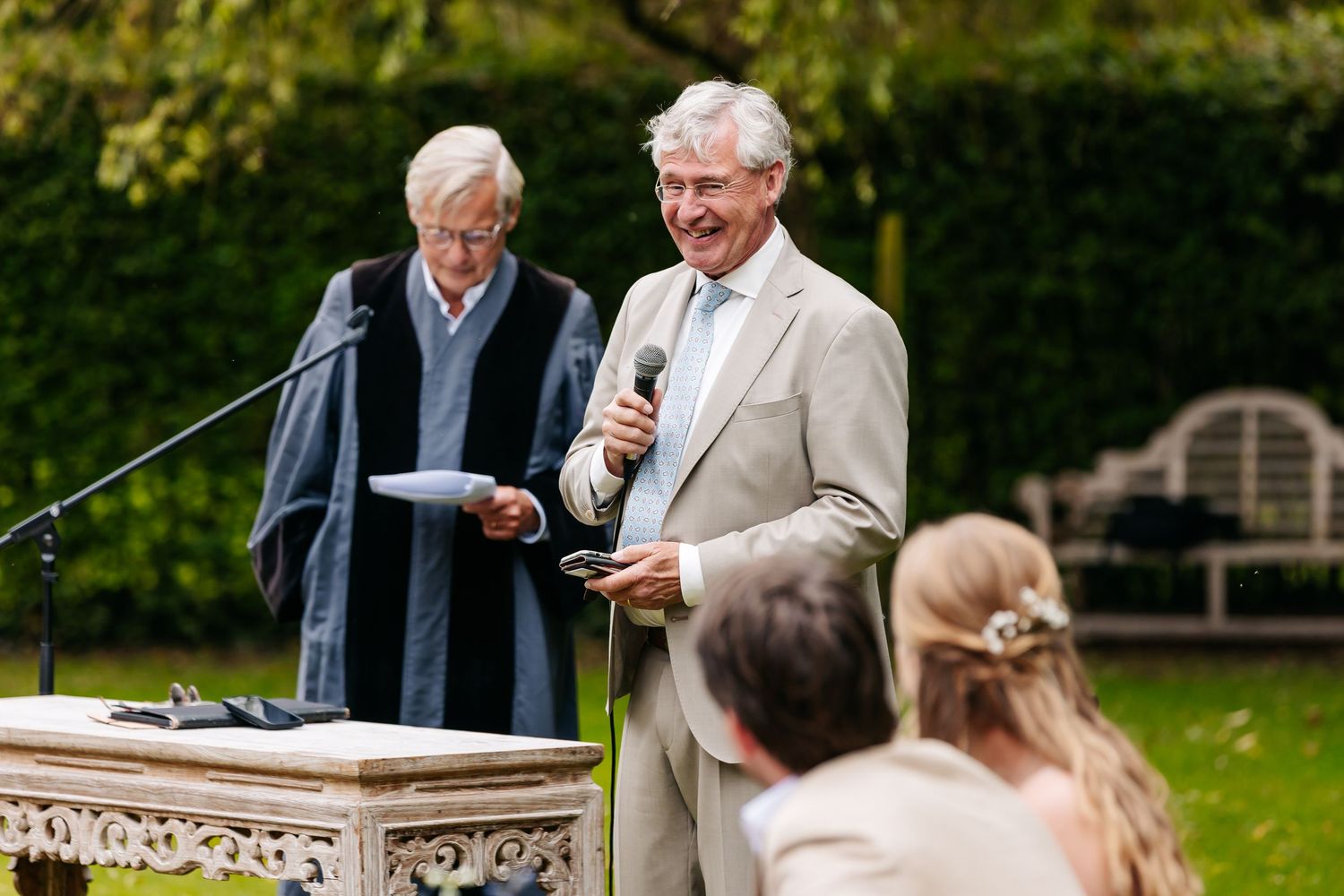 Man giving a heartfelt speech at an outdoor wedding ceremony beside an officiant at the altar.