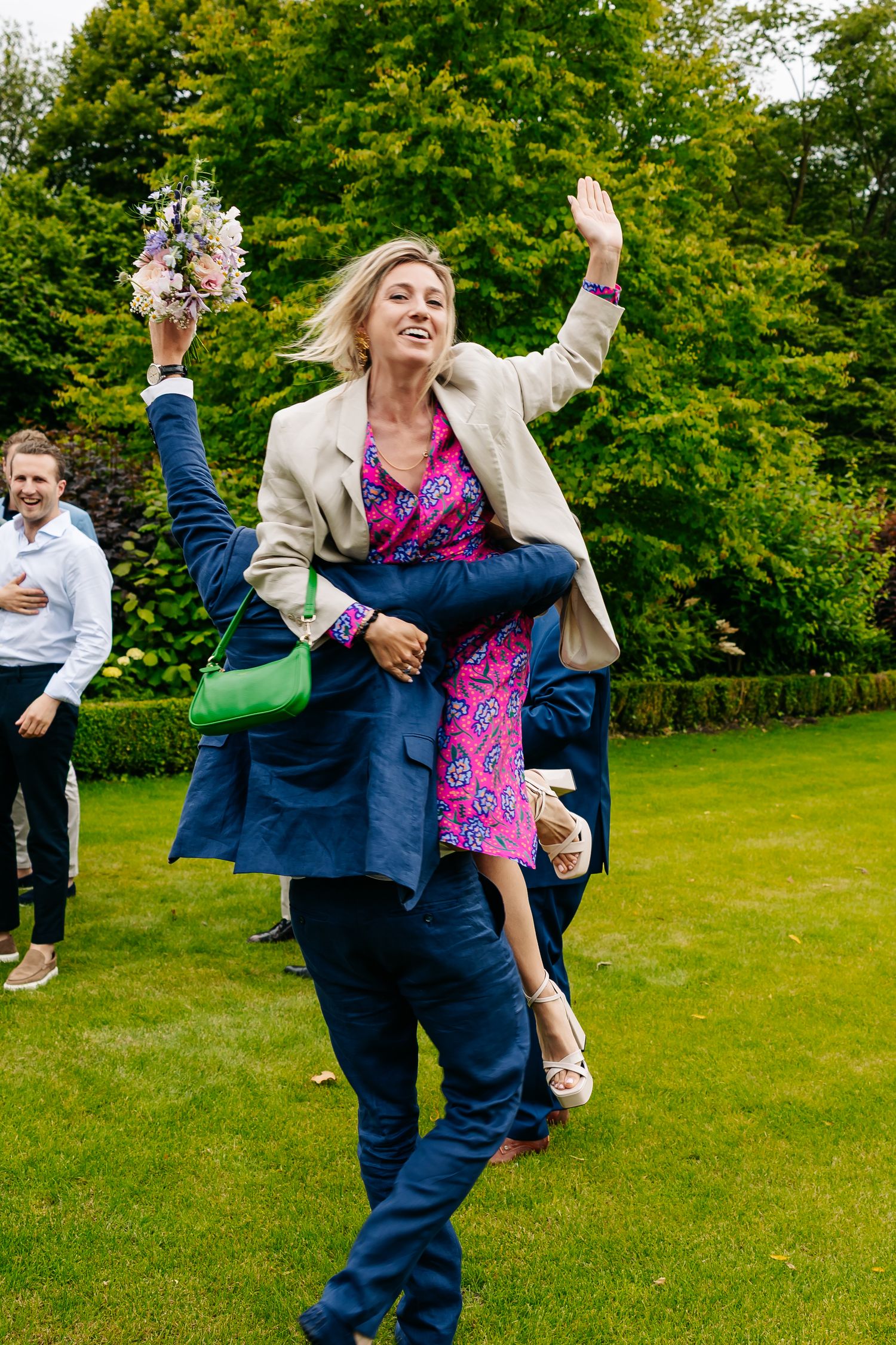 Joyful woman in pink floral dress being carried by a man in navy suit while holding a bouquet at a wedding.