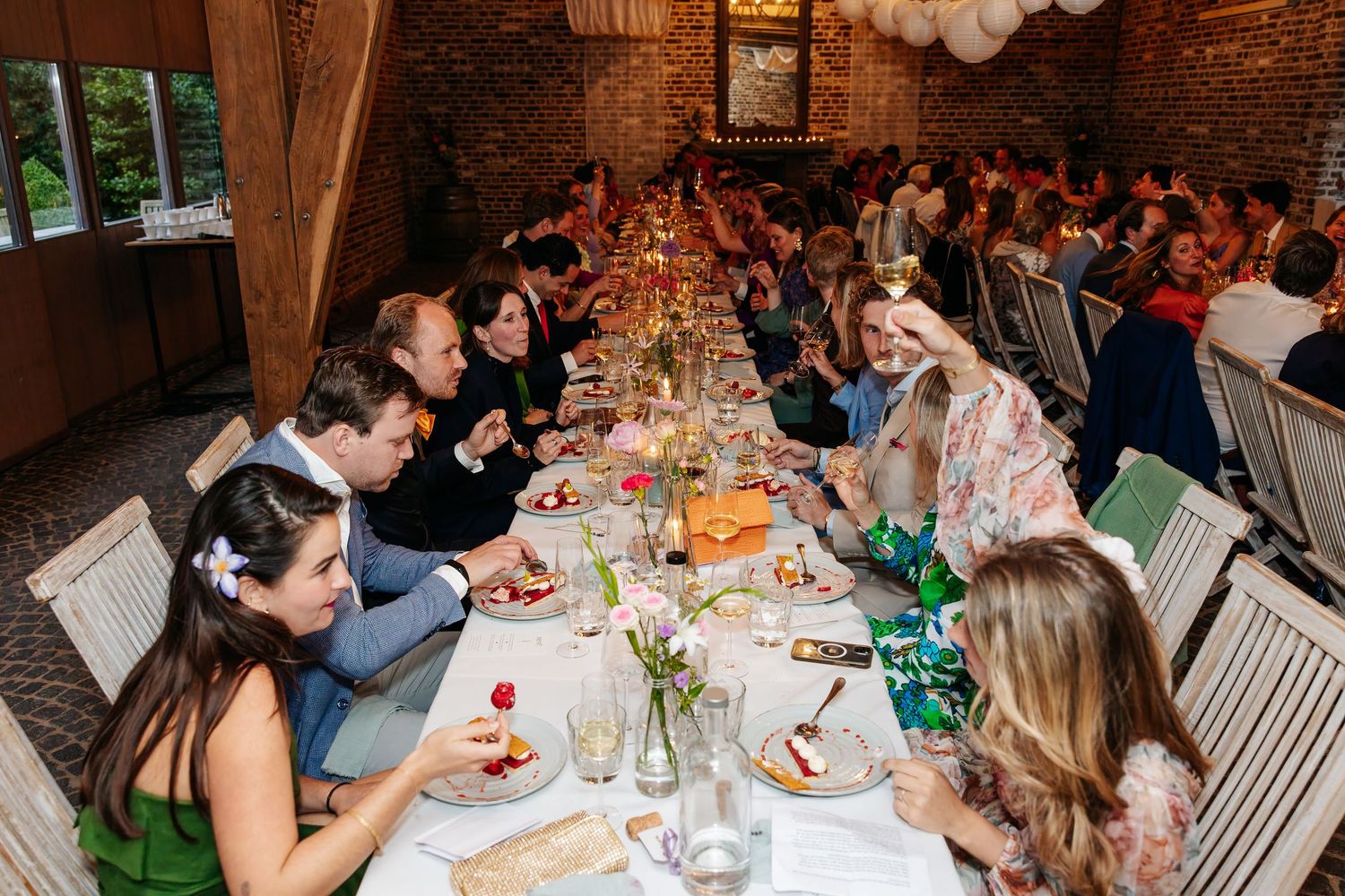 Wedding guests enjoying dinner at long decorated table in rustic brick venue with warm lighting.