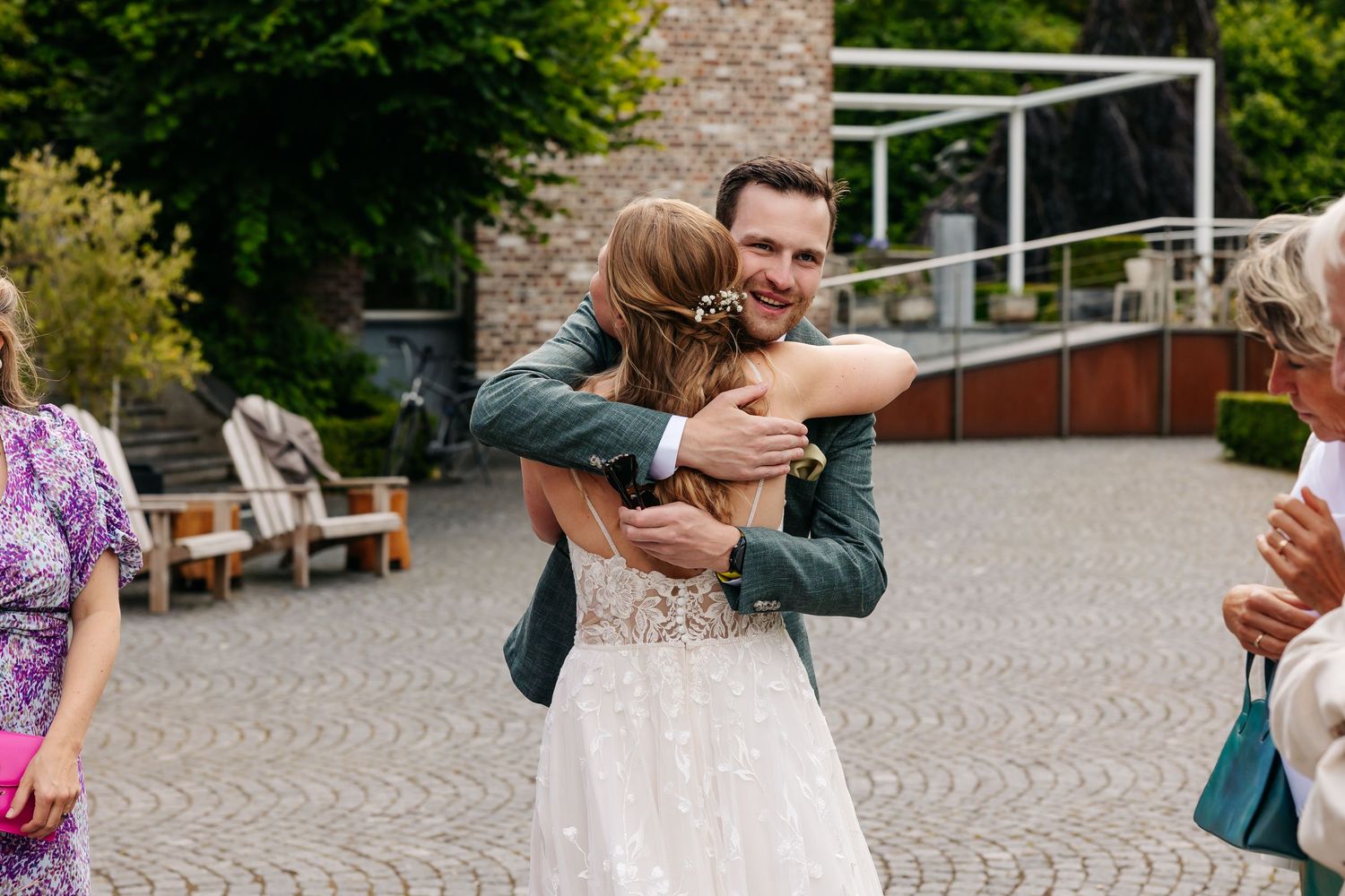 Smiling groom in teal suit embracing bride in white lace dress at outdoor wedding venue.