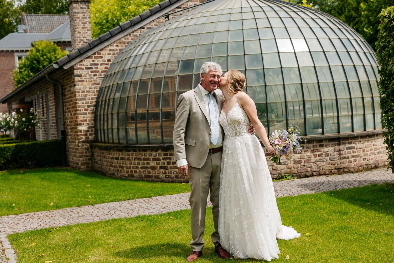 Bride and older gentleman share a tender moment outside a glass greenhouse on a sunny wedding day.
