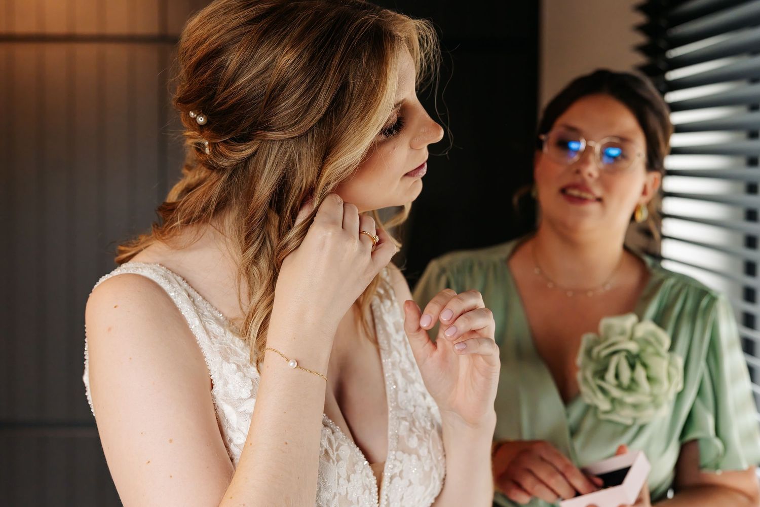 Bride in white lace gown touches her cheek as bridesmaid in green dress looks on during wedding preparations.