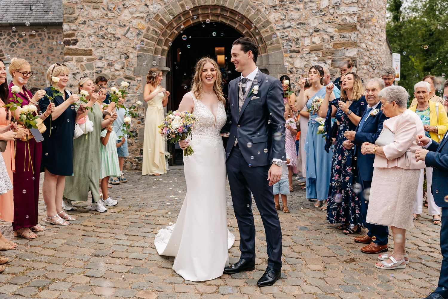 Smiling newlyweds walk through stone church entrance surrounded by colorfully dressed wedding guests.