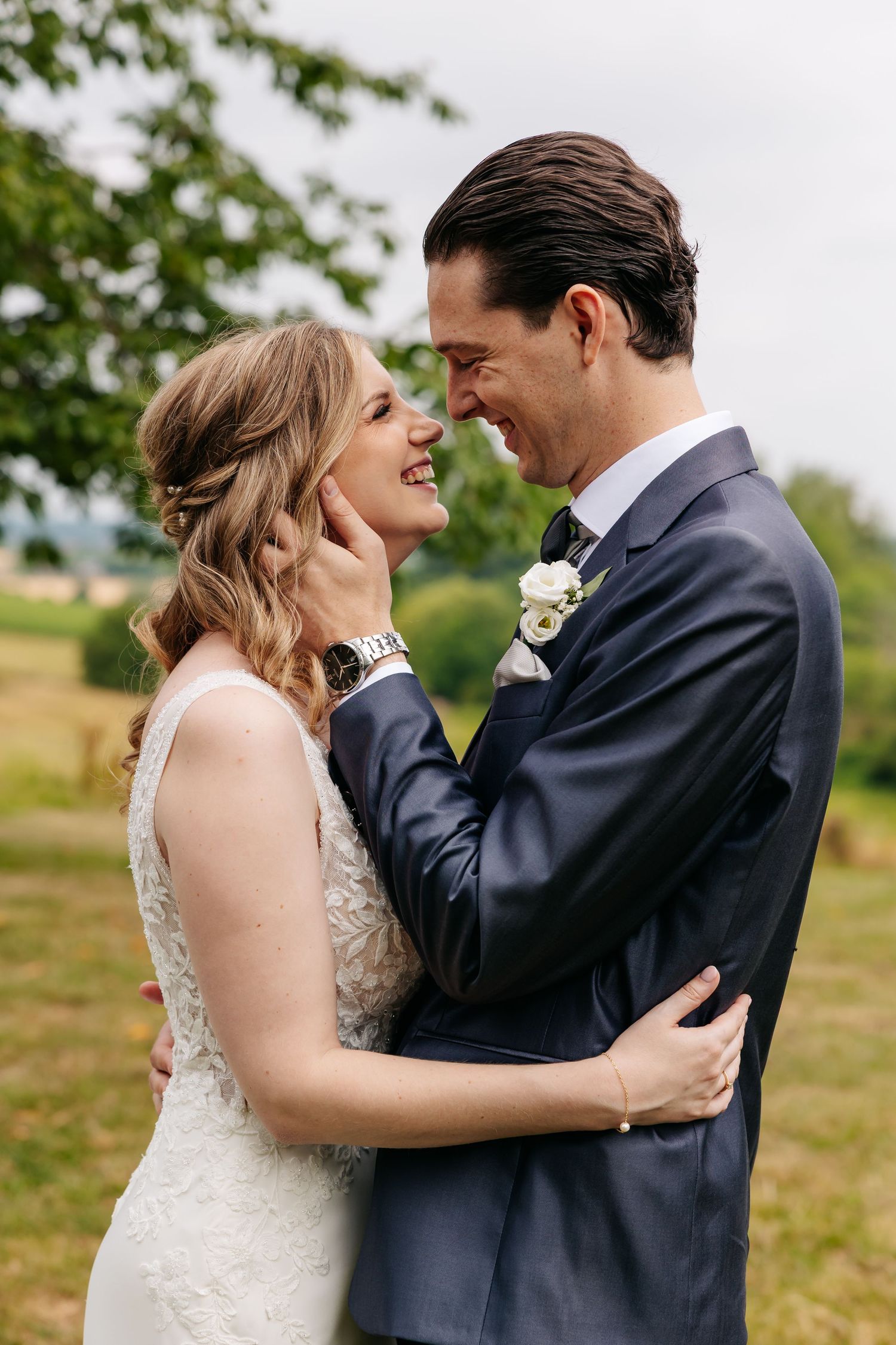 Groom tenderly cupping the bride's face as they laugh together outdoors on their wedding day.