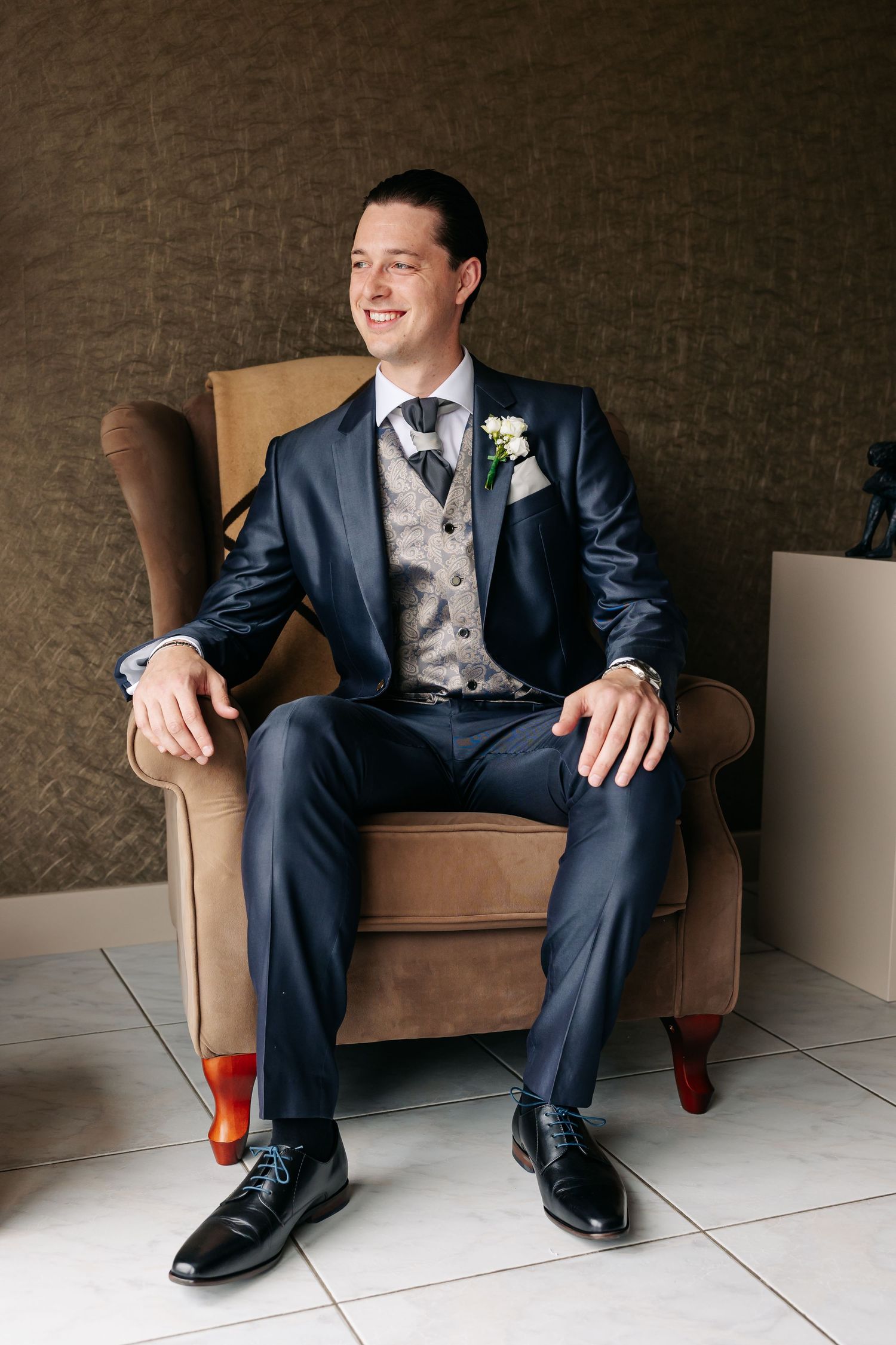Handsome groom in navy three-piece suit sitting relaxed in brown armchair smiling.