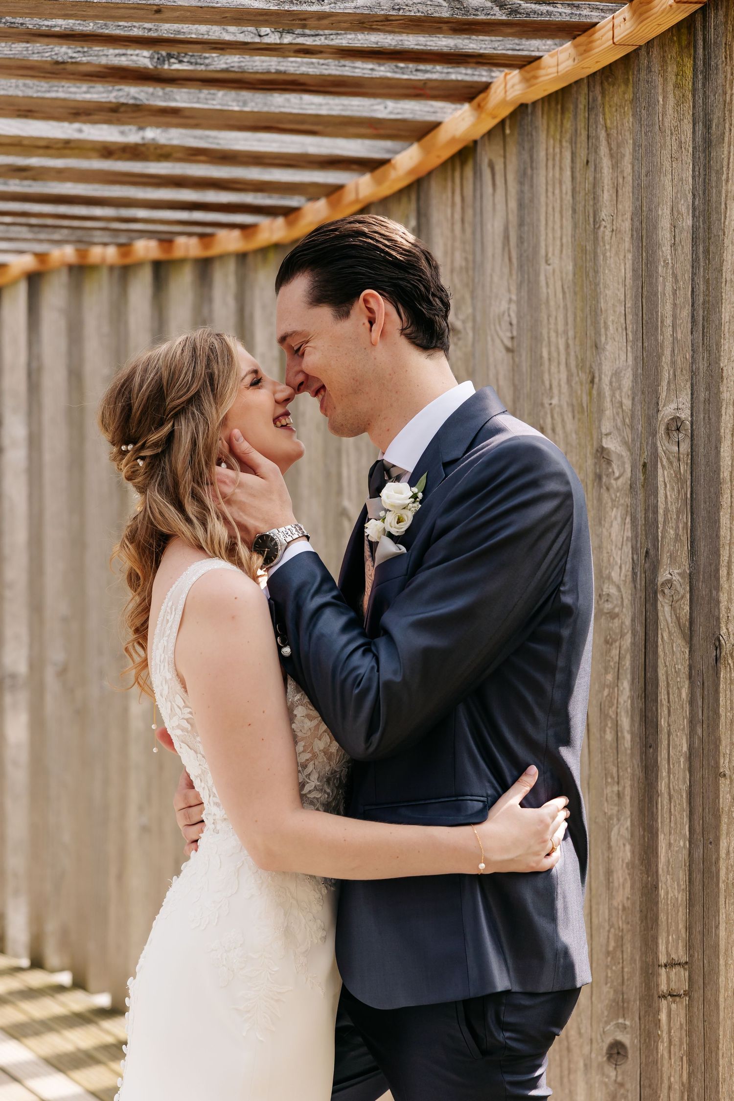 Smiling bride and groom touch foreheads lovingly beside rustic wooden fence on wedding day.