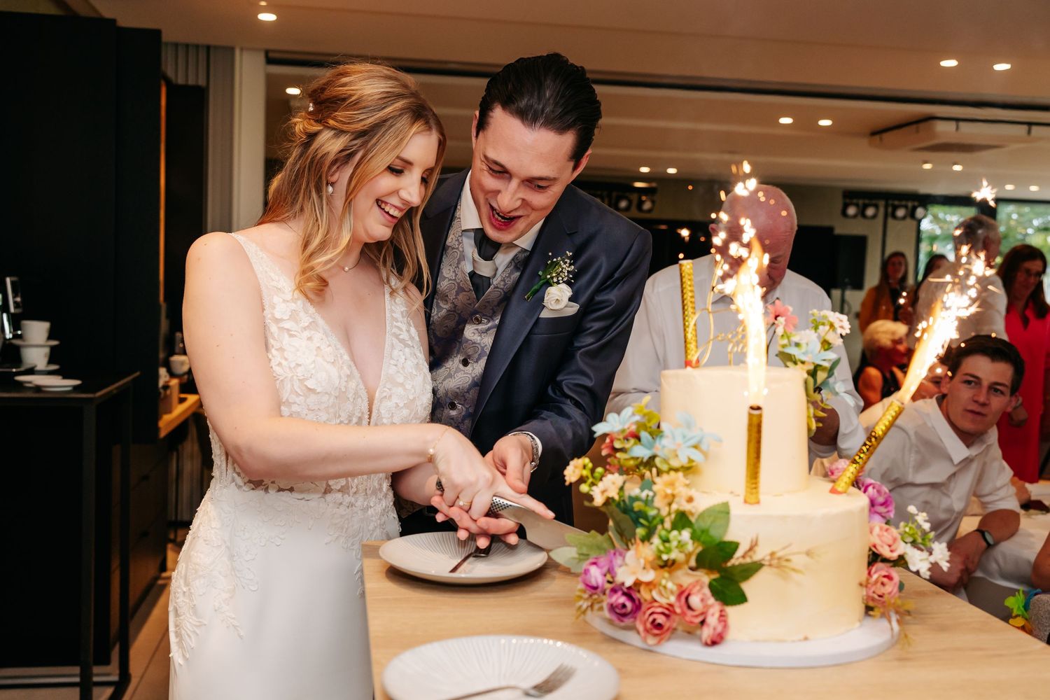 Bride and groom cutting a white wedding cake decorated with sparklers and colorful flowers.