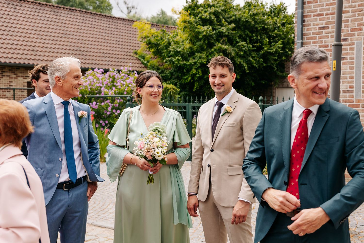 Smiling bride in sage green dress holds bouquet with groom in tan suit at outdoor wedding reception.
