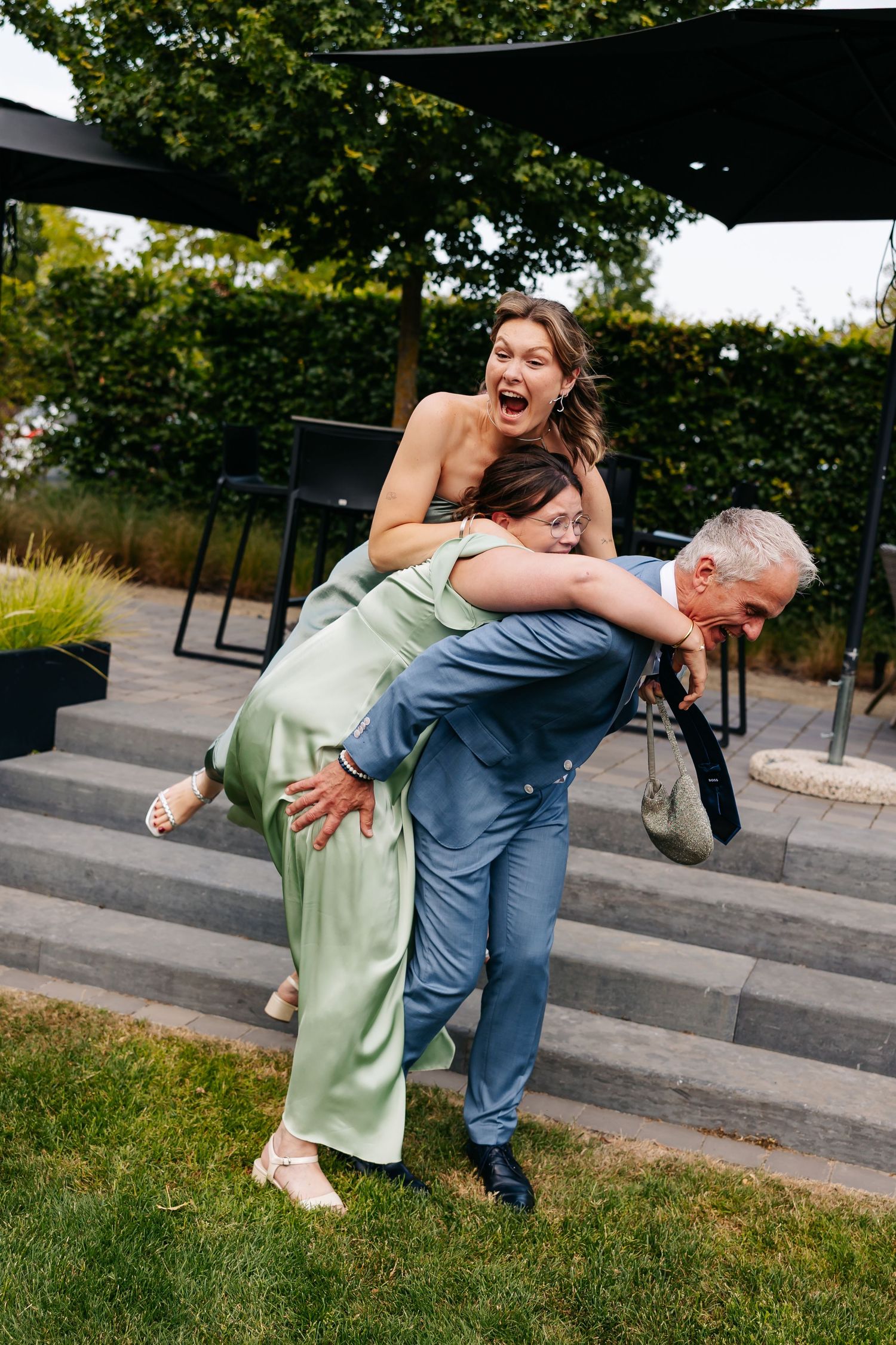 Three wedding guests laugh as a man in a blue suit lifts two women in green dresses outdoors.