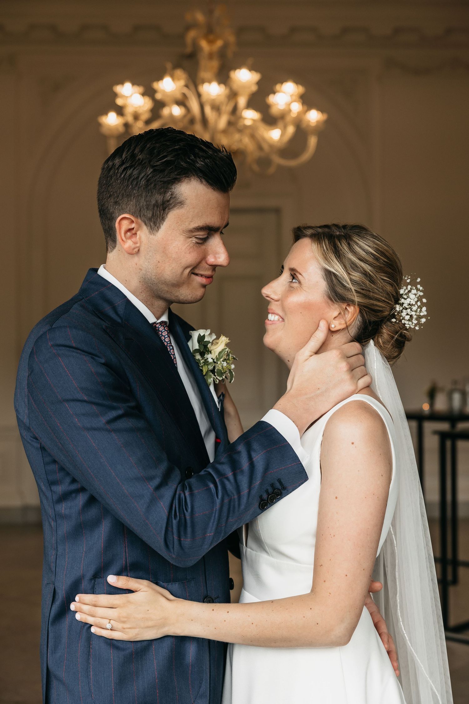 Bride and groom sharing tender moment under chandelier at indoor wedding ceremony.