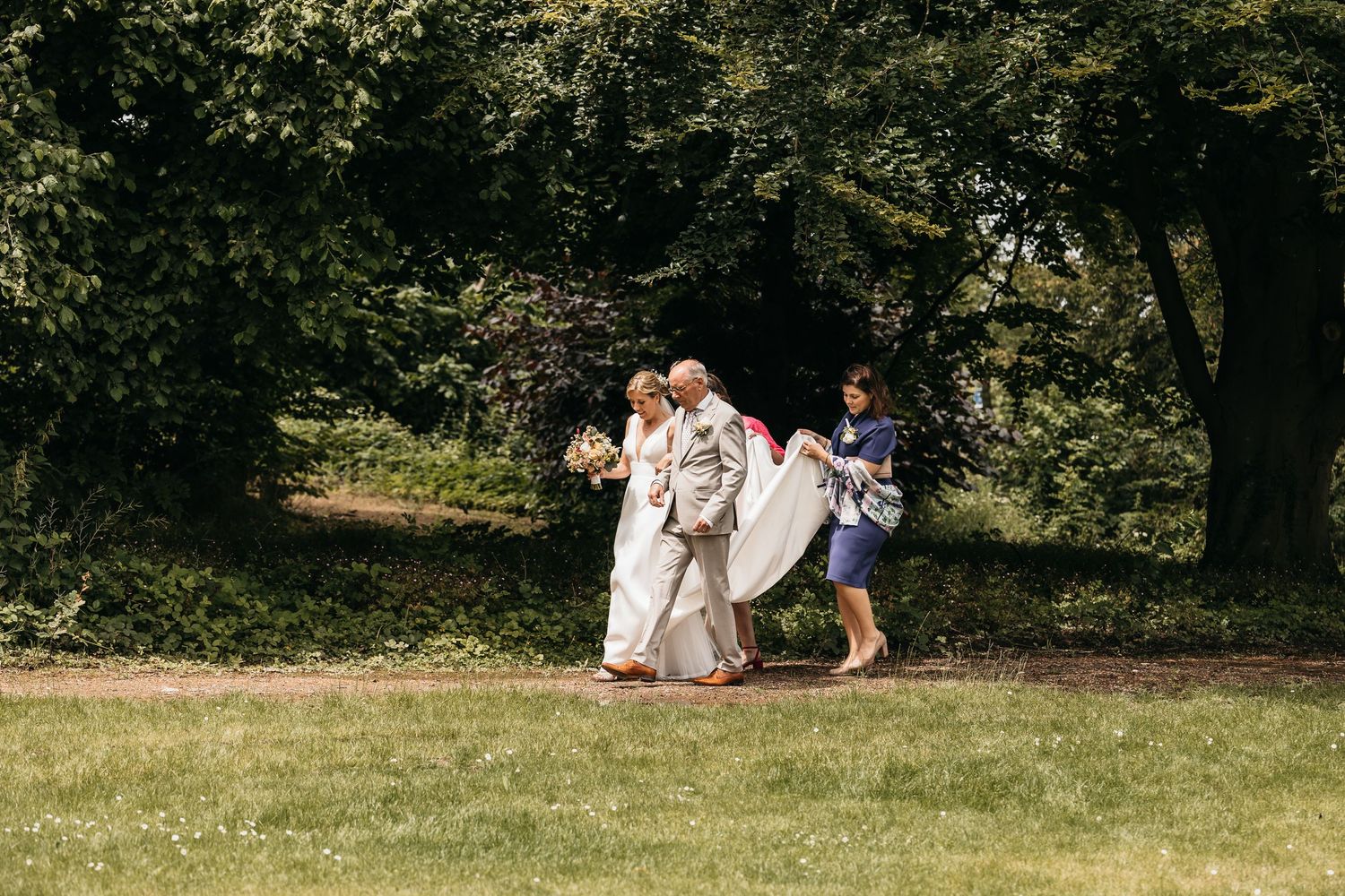 Bride in white gown walks through lush green garden accompanied by two attendants carrying her train.