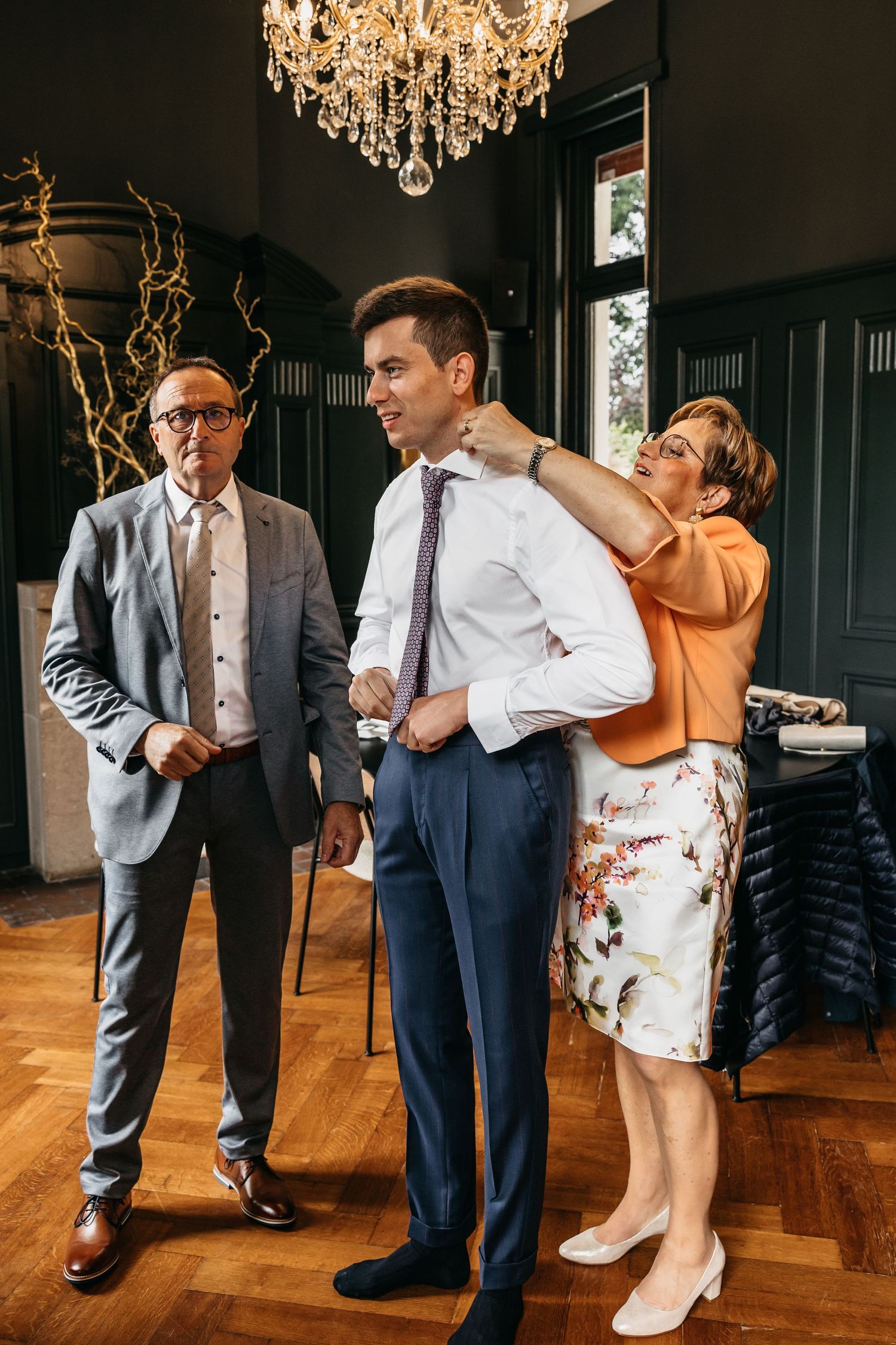 Parents helping groom adjust his tie and collar under a crystal chandelier before wedding.