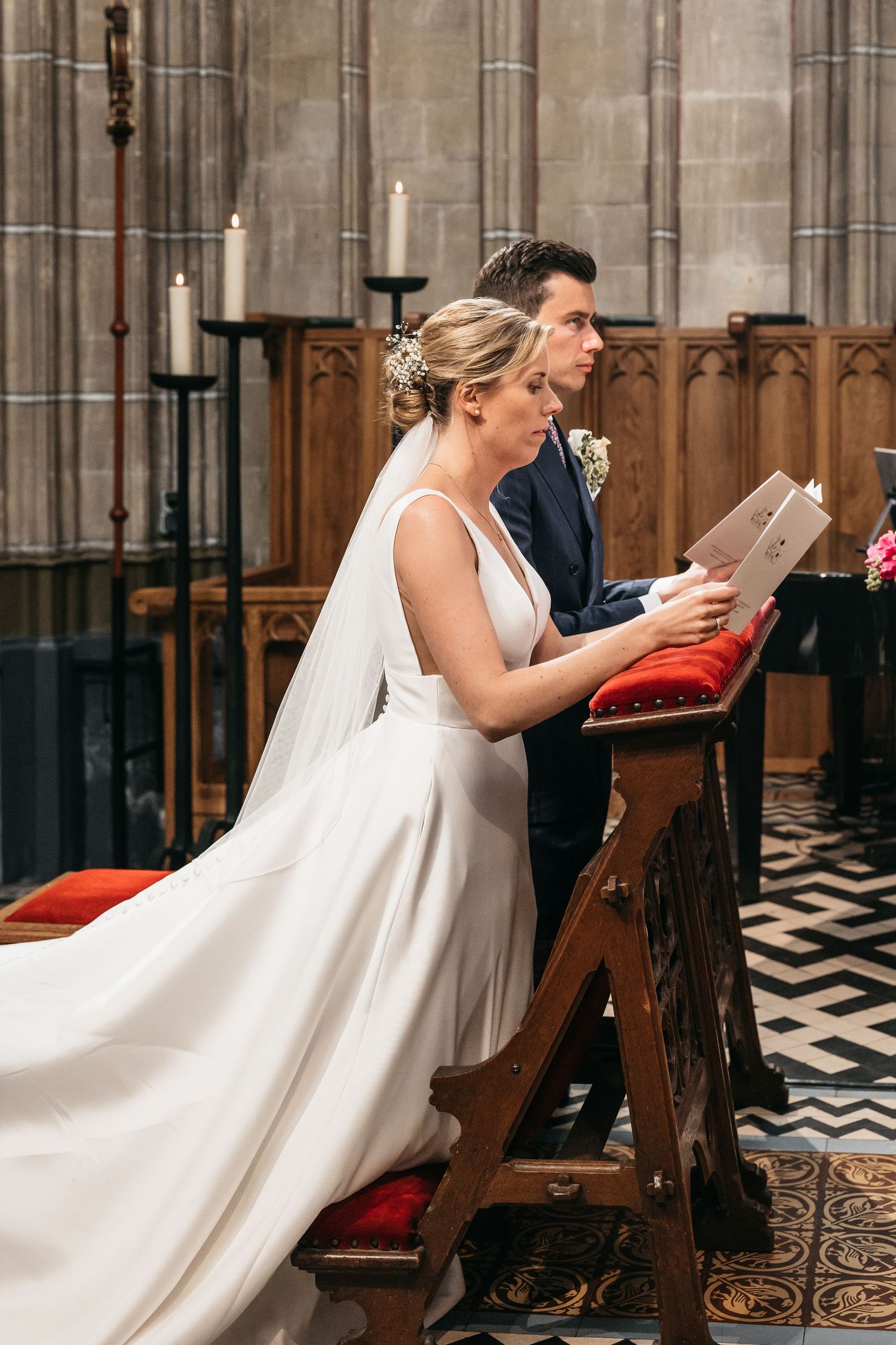 Bride in white gown and groom kneel at wooden prie-dieu reading during church wedding ceremony.