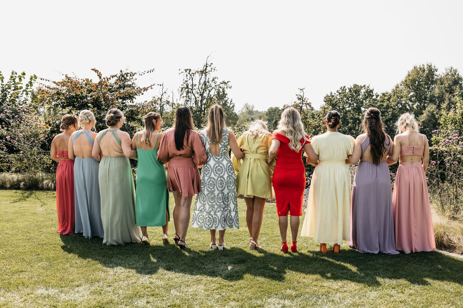 Bridesmaids in colorful mismatched dresses viewed from behind standing together in sunny garden.