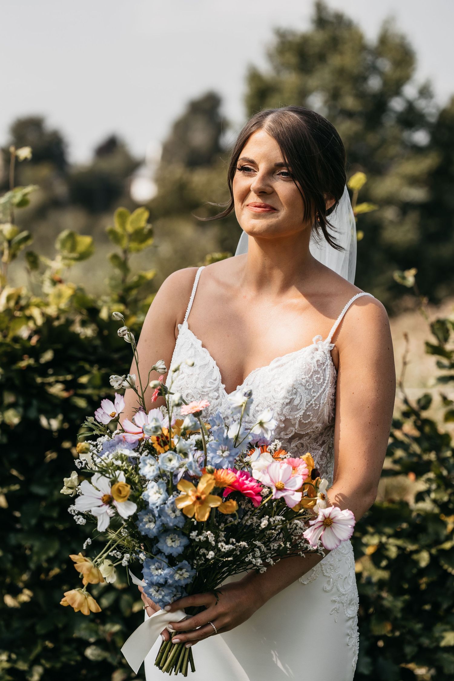 Smiling bride in white lace dress holding vibrant mixed flower bouquet outdoors.