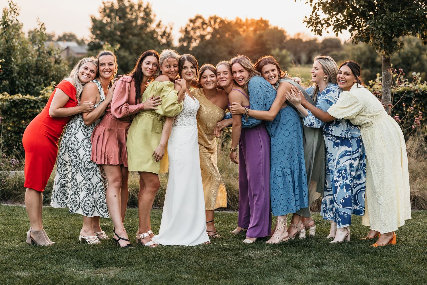 Bride surrounded by smiling female guests in vibrant dresses hugging at outdoor wedding.