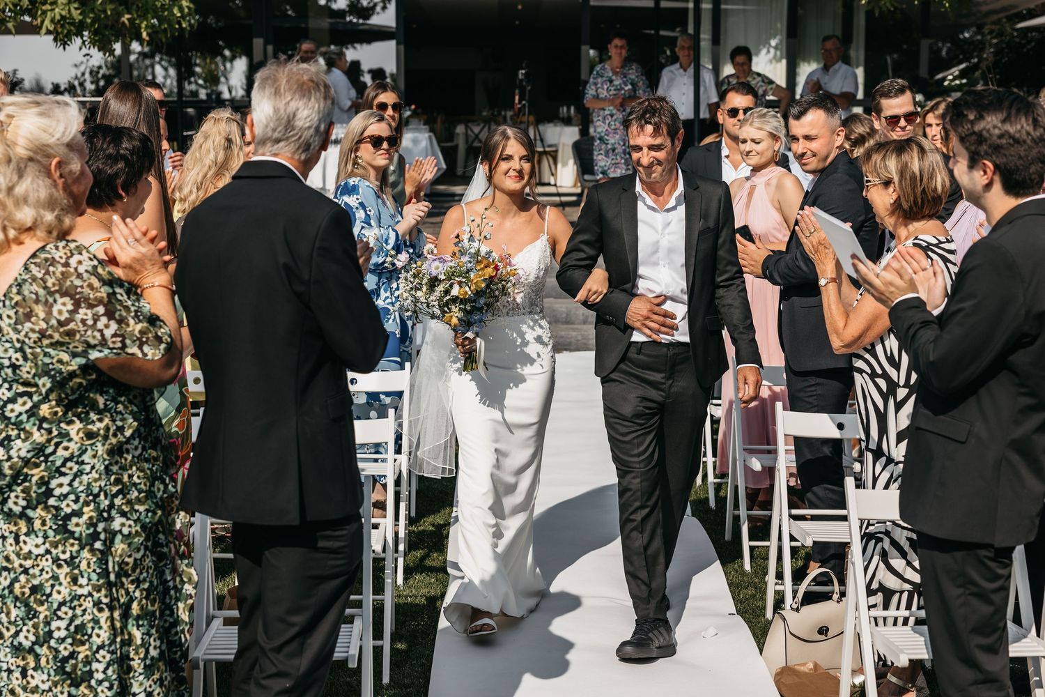 Smiling bride holding wildflower bouquet walks with groom down sunlit outdoor wedding aisle.