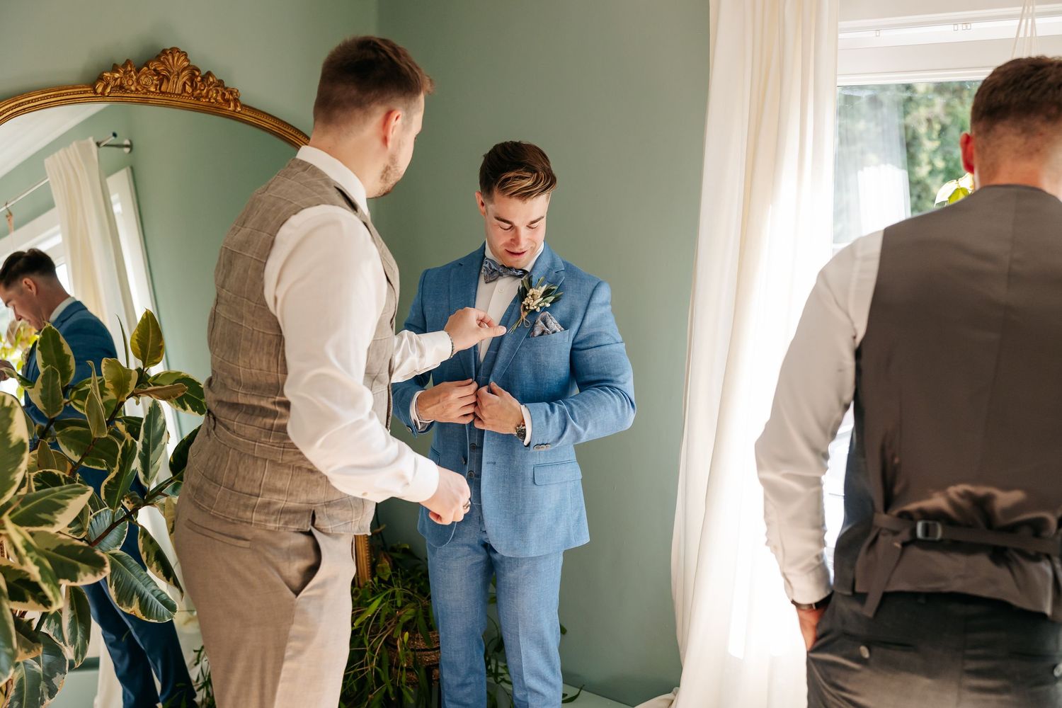 Groomsmen helping groom button up light blue suit jacket before wedding ceremony.